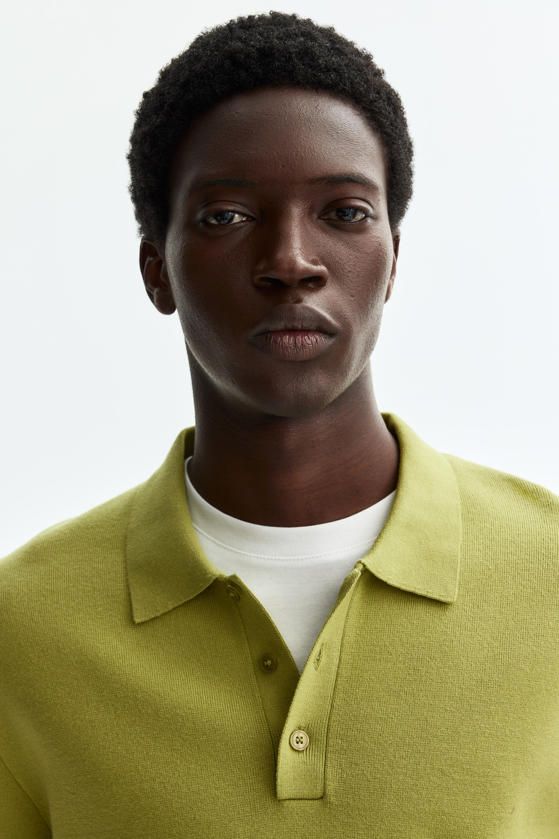 View larger image: "A lime green knit polo shirt featuring a classic collar and a two-button placket with light brown buttons. It is worn over a white crew-neck t-shirt."