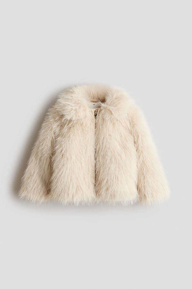 A cream-coloured faux fur jacket is shown from the front, showcasing a plush, fluffy texture and a rounded collar. It features a front zip fastening with a small gold-tone, heart-shaped zip pull and long, straight-cut sleeves. - 1
