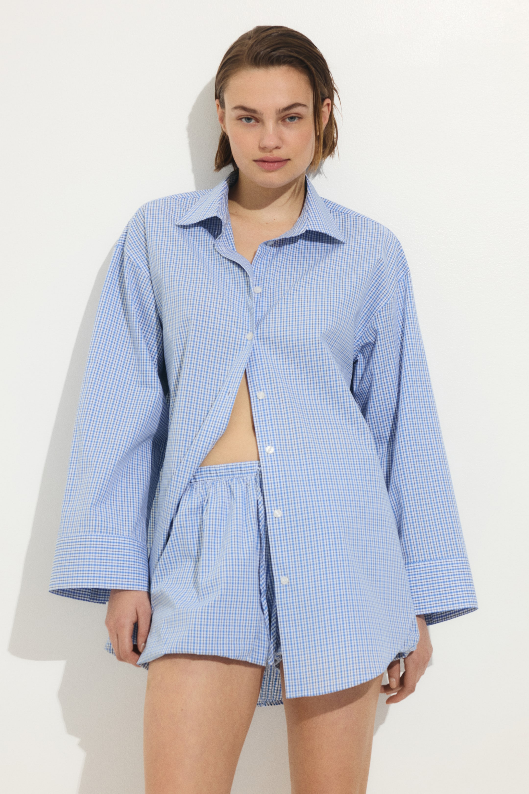 View larger image: A blue and white gingham sleepwear set includes an oversized long-sleeved shirt with a classic collar and button-up front, styled unbuttoned from the chest. The matching shorts feature an elastic waistband and a loose fit, with the set facing forward.