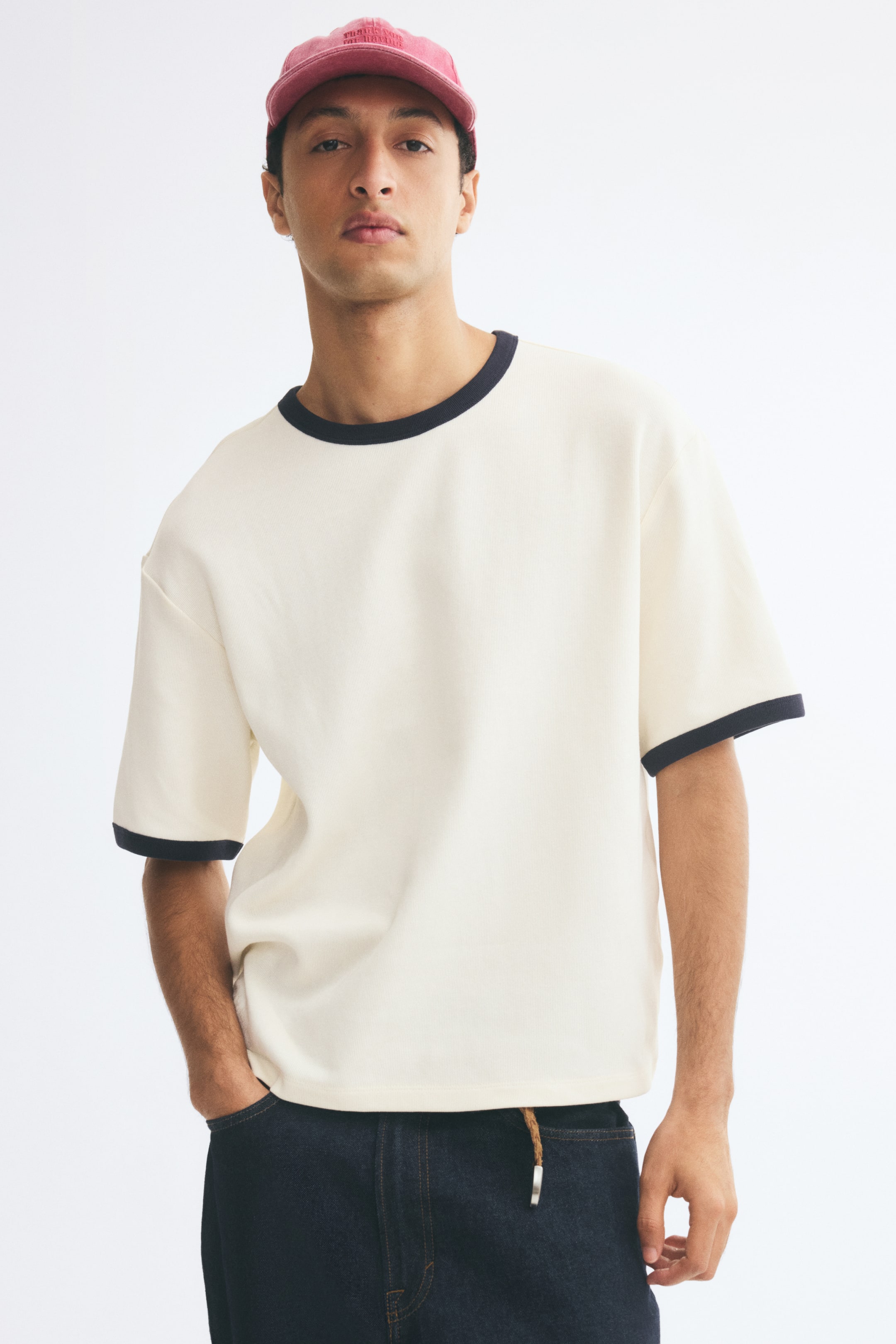 View larger image: A cream-colored, relaxed-fit T-shirt featuring dark contrast trim along the crew neckline and short sleeve cuffs is worn by the model. The T-shirt has dropped shoulders and is styled with dark denim jeans.