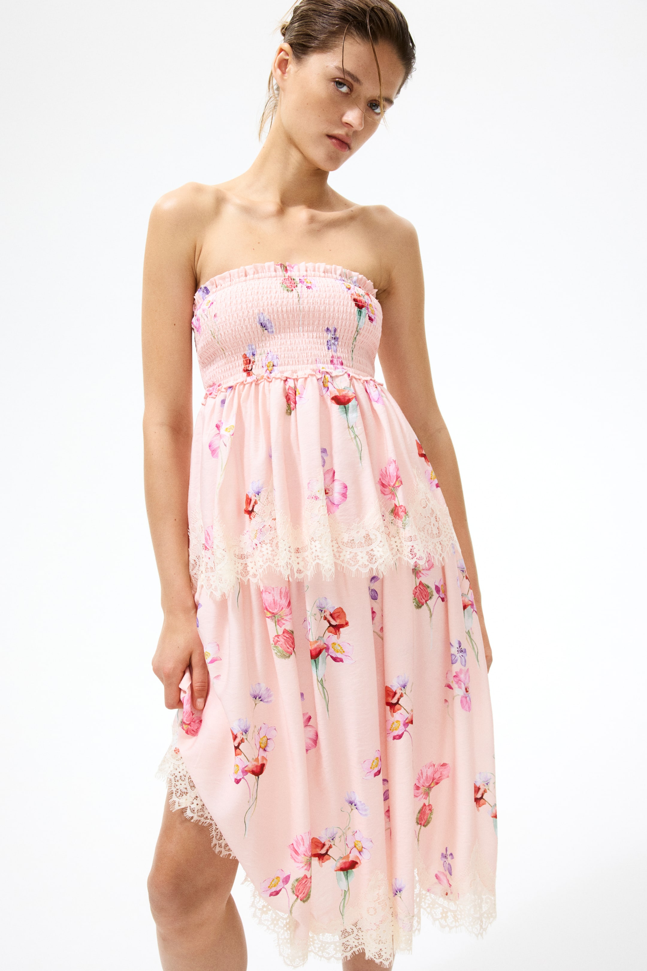 View larger image: The model is wearing a calf-length bandeau dress in a pink floral print. The dress features a fitted, smocked top section with narrow frill trims and a flared skirt showcasing a wide, lace-trimmed tier with gathers below the bust for added volume, along with a lace trim at the irregular hem.