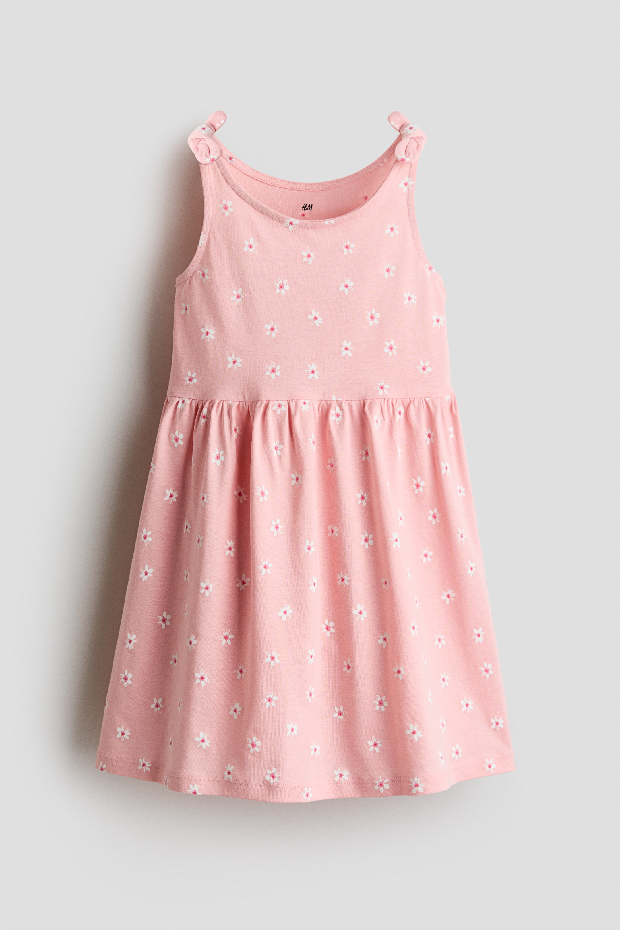 View larger image: The dress is a pink, sleeveless piece showcasing a white and pink floral pattern throughout. It features thin shoulder straps tied into bows and a gathered seam at the waist, creating a flared skirt.