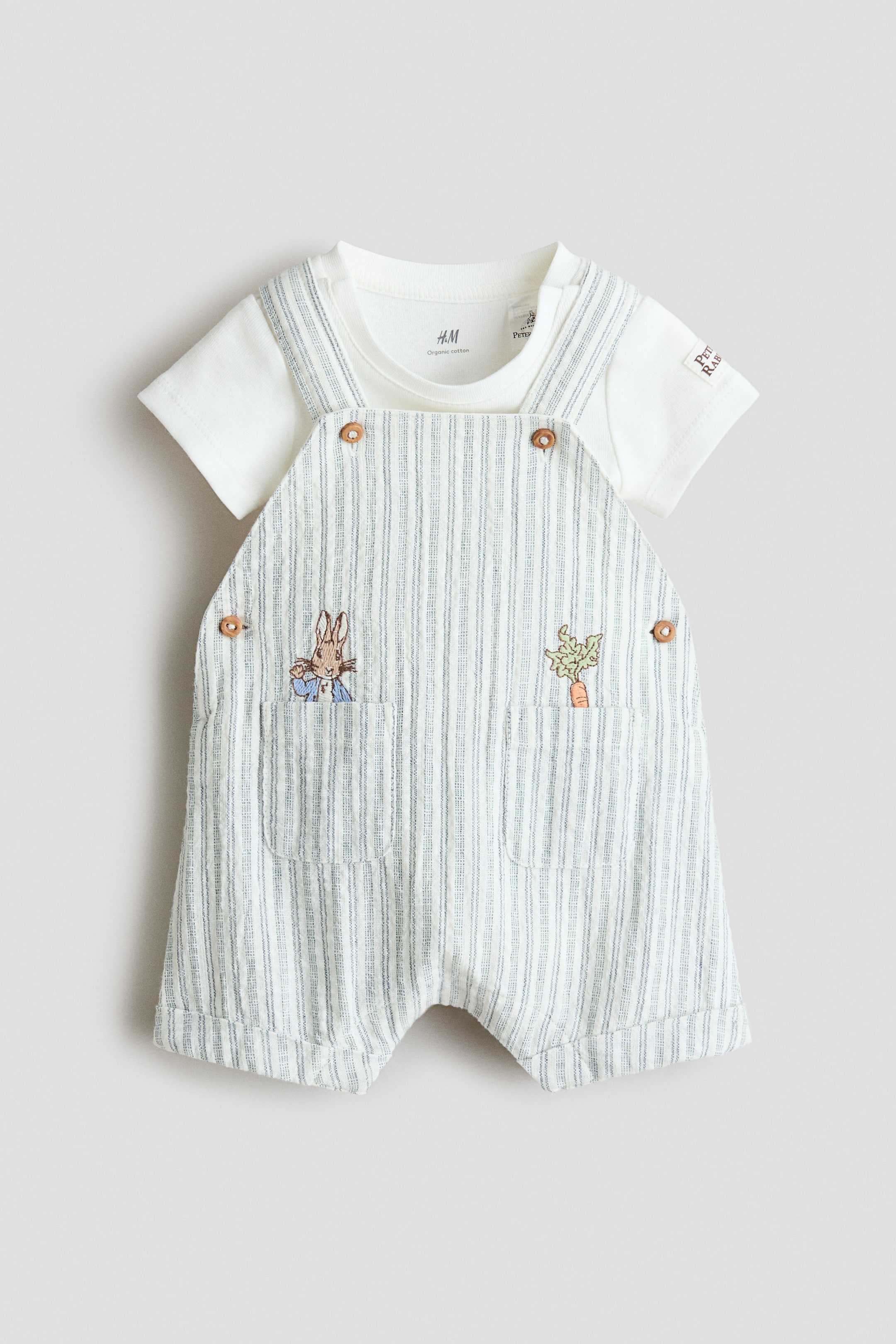 Grotere afbeelding bekijken: Een babyset met korte overall en t-shirt wordt frontaal gepresenteerd. Een wit t-shirt met korte mouwen wordt onder de korte overall gedragen, die een witte basis heeft met dunne verticale blauwe strepen, twee voorzakken met geborduurde Peter Rabbit- en wortelontwerpen, en houten knopen op de schouderbanden.