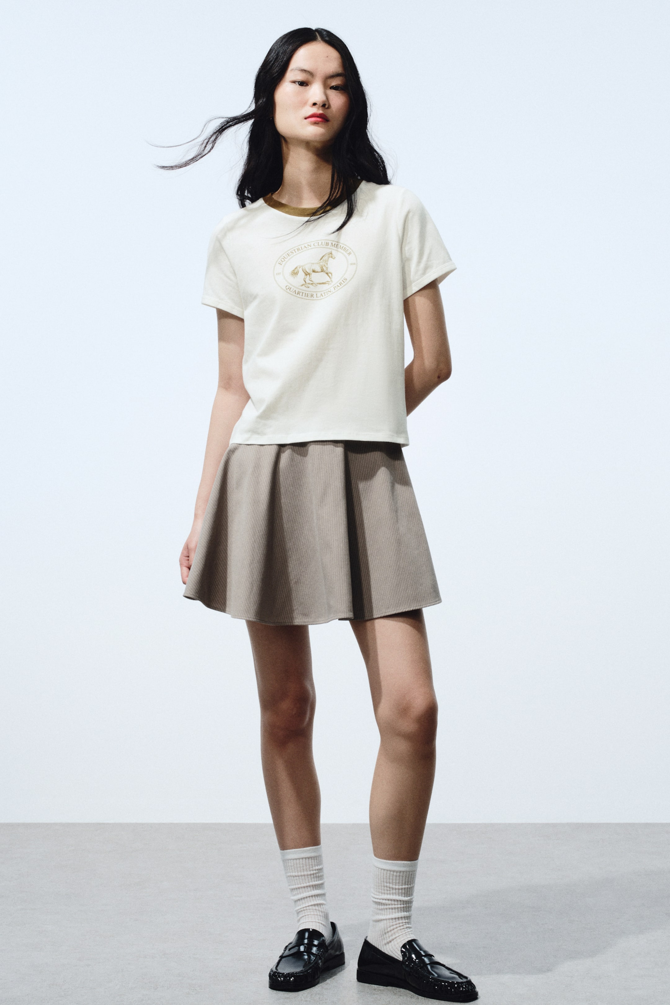 View larger image: An off-white, short-sleeved T-shirt features a golden horse graphic set within a circular emblem on the front, with a contrasting brown trim on the crew neck. A beige mini skirt with subtle vertical ribbing complements the T-shirt, flaring out from the waist.