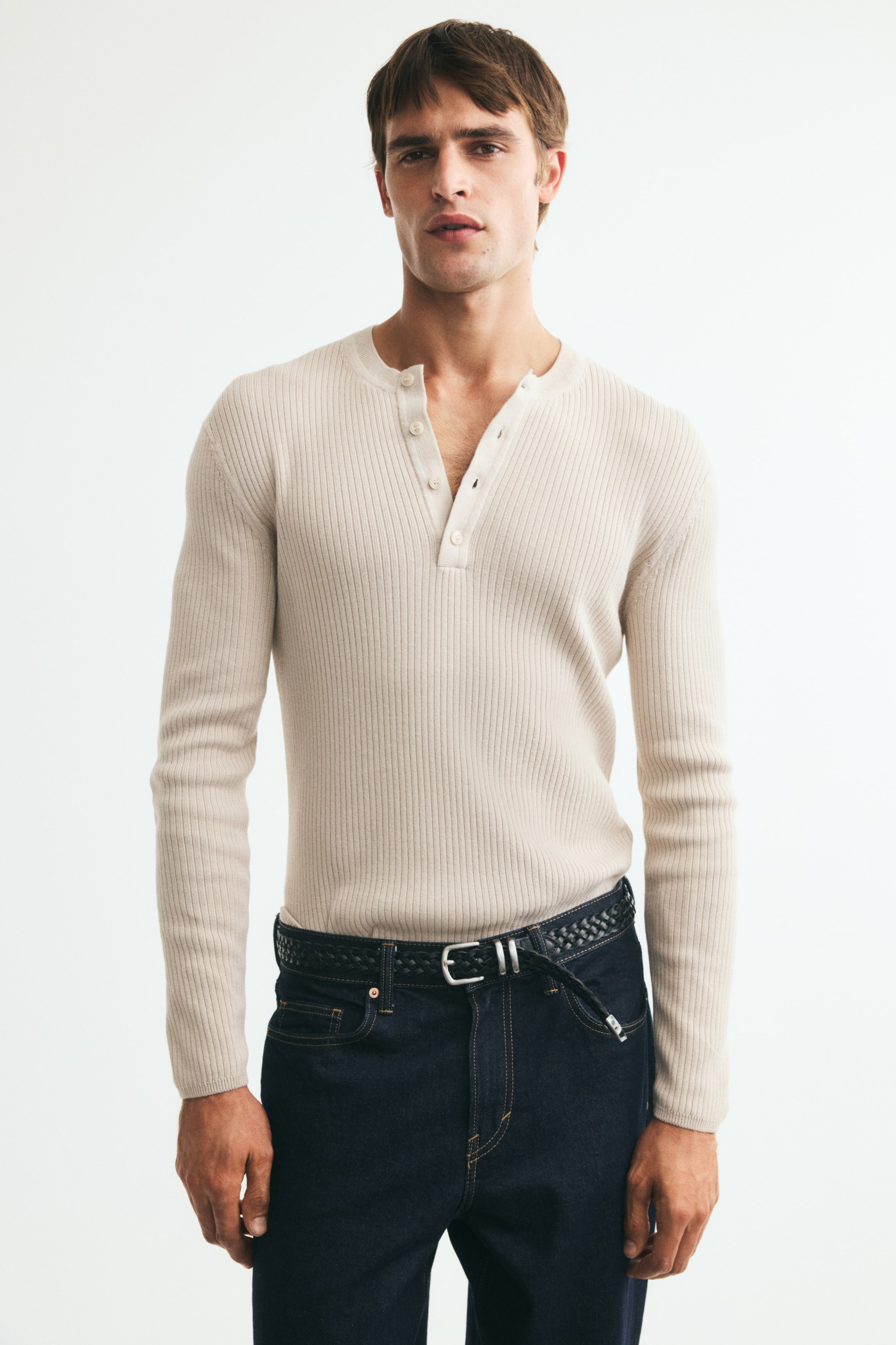 View larger image: A cream-coloured, ribbed-knit long-sleeve top is being worn by the model, featuring a slim fit, a round neckline, and an open button placket. The top is tucked into dark blue denim jeans styled with a black braided belt.