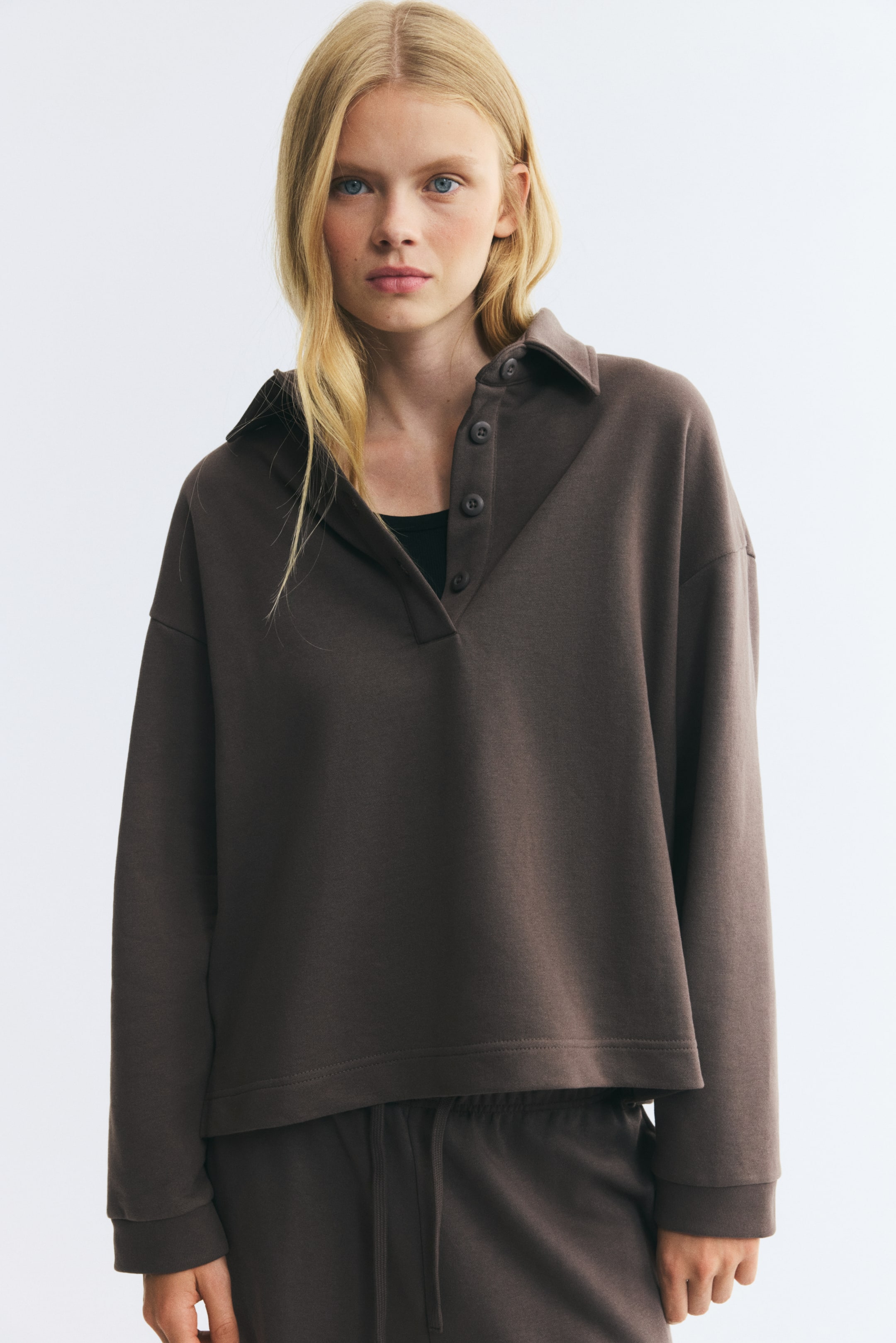 Ver imagem maior: A modelo está a vestir uma camisola castanha-escura em tecido de sweat leve, com gola, carcela de botões parcialmente aberta e ombros descaídos. A camisola, sobreposta a um top preto e combinada com umas sweatpants a condizer, tem mangas compridas com punhos canelados e bainha de corte reto.