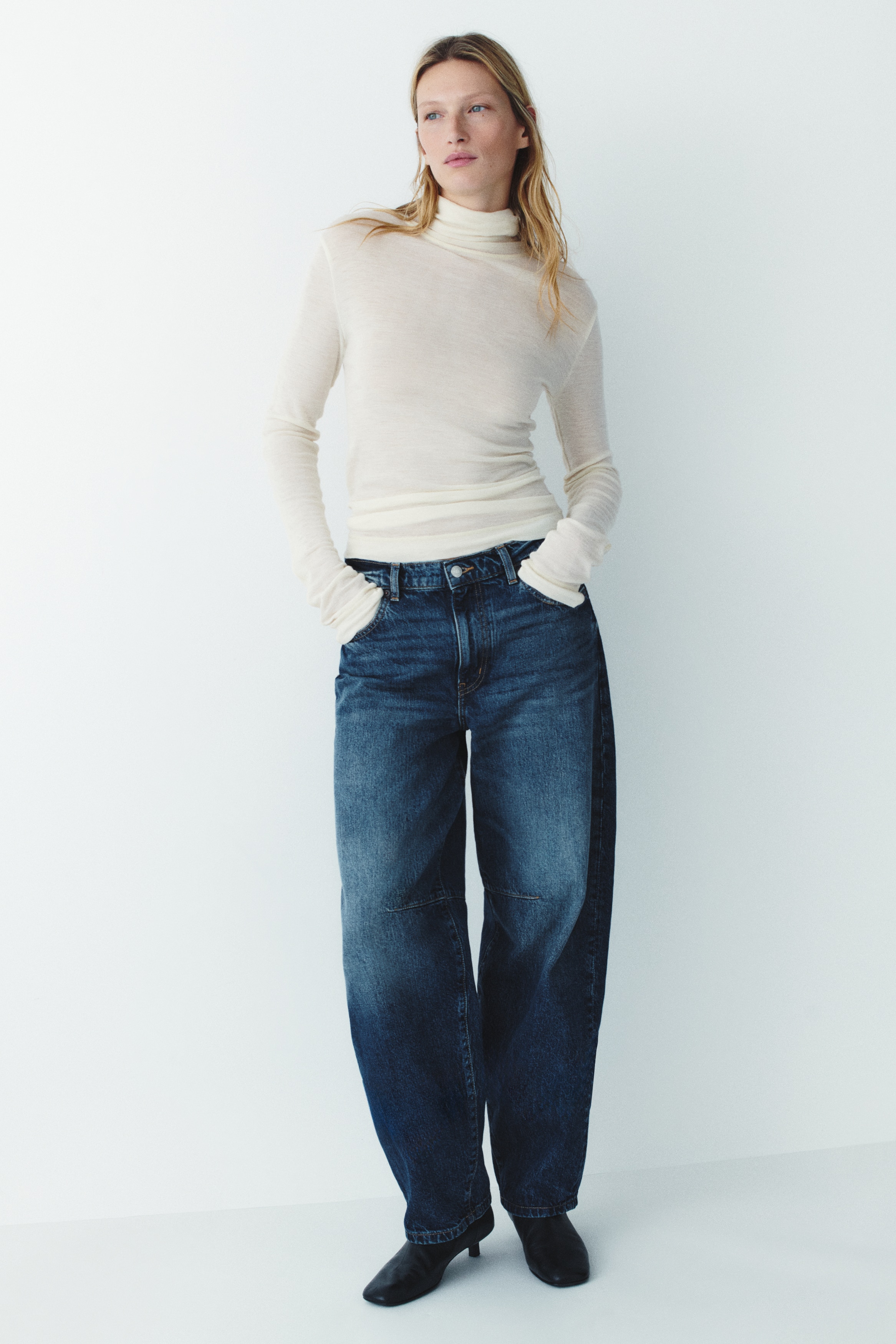 These blue barrel-leg jeans are shown from the front, featuring a high-rise waist with belt loops and classic five-pocket styling. They have a curved, tapered silhouette with a faded wash down the centre of the legs and distinctive horizontal seam details at the knees.