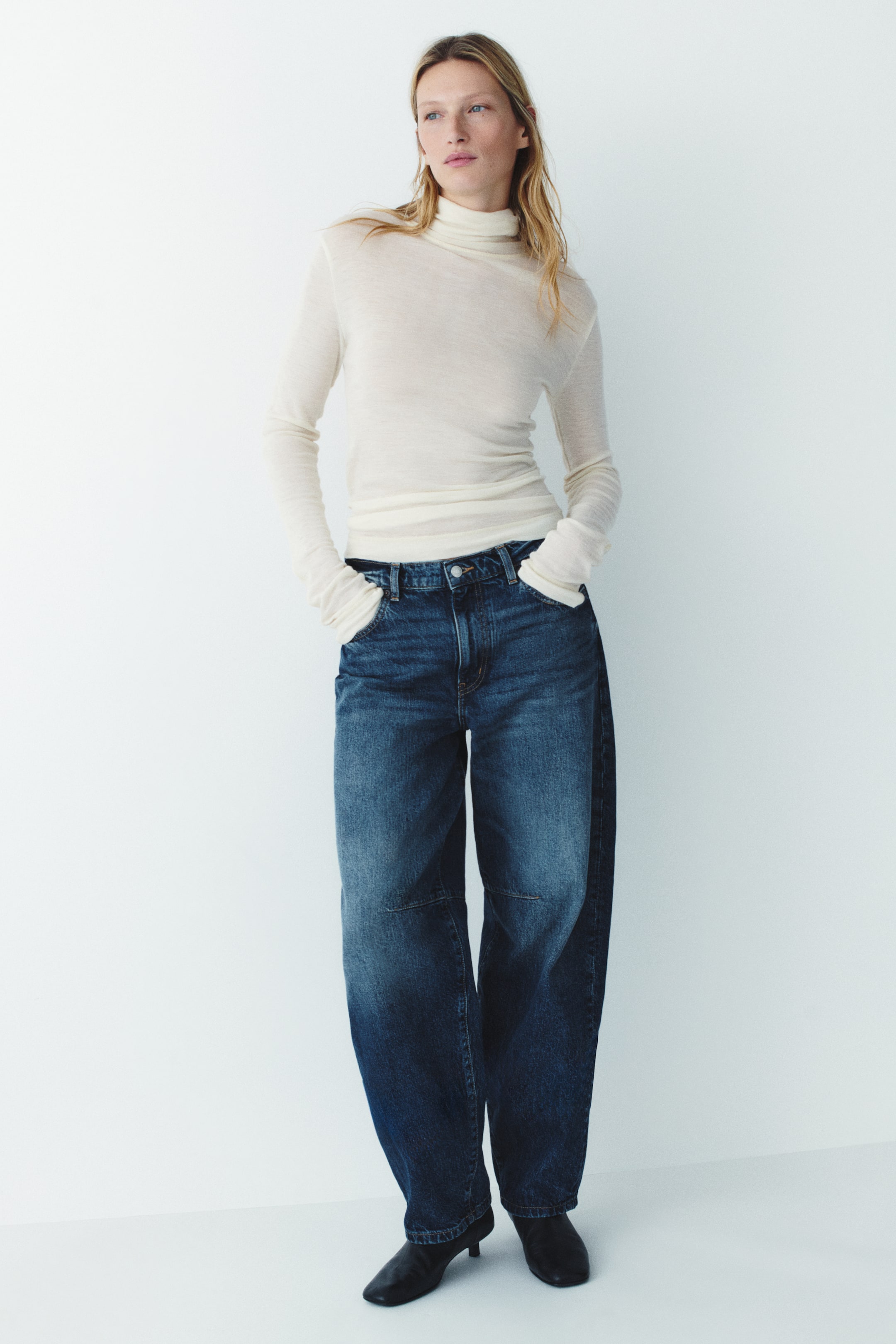 View larger image: Dark wash blue denim jeans featuring a curved, voluminous leg silhouette and knee darts are being worn by the model. The model is standing with hands resting in the pockets, styling the regular-waist jeans with a cream-coloured roll-neck top and black shoes.