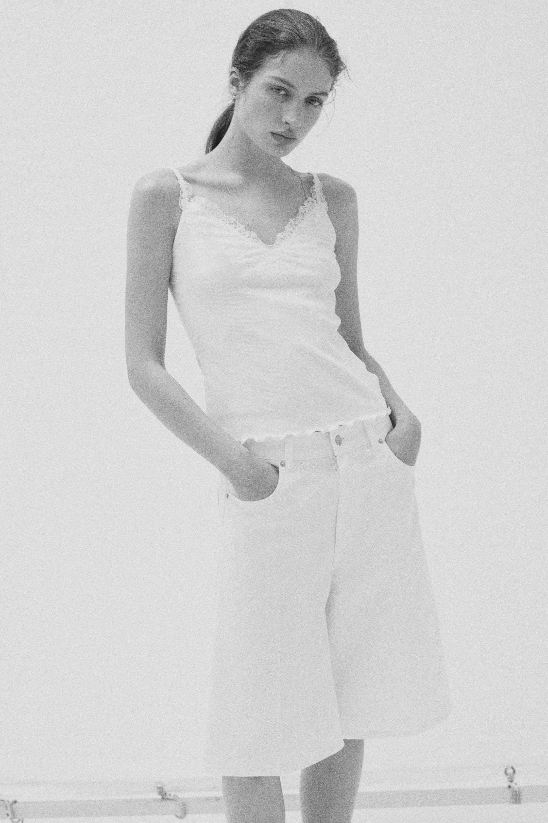 Größeres Bild ansehen: Ein weißes Camisole mit einem V-Ausschnitt, der mit Spitze verziert ist, dünnen Trägern und einem Saum im "Lettuce-Edge"-Stil wird mit weißen, hoch taillierten Shorts mit weitem Bein kombiniert, die über Fronttaschen verfügen und bis über das Knie reichen.