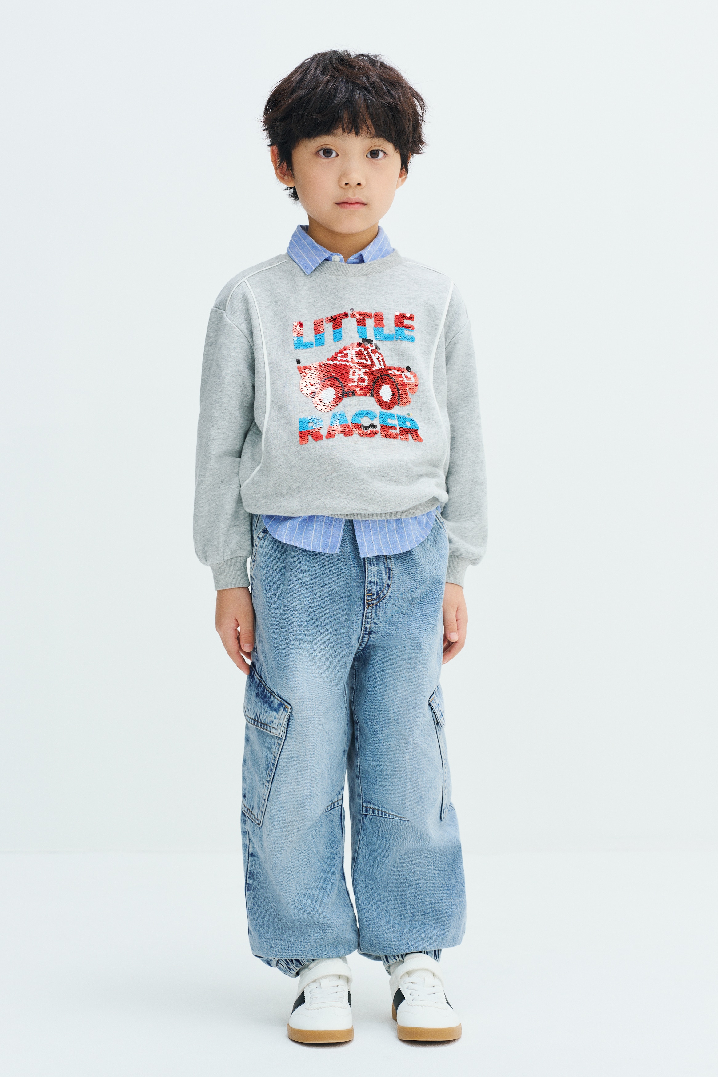 Light blue washed denim joggers are shown from the front, featuring a gathered elasticated waistband and a faux fly. The balloon-leg trousers feature diagonal side pockets, mid-leg patch pockets with flaps, and gathered elasticated cuffs at the hems.