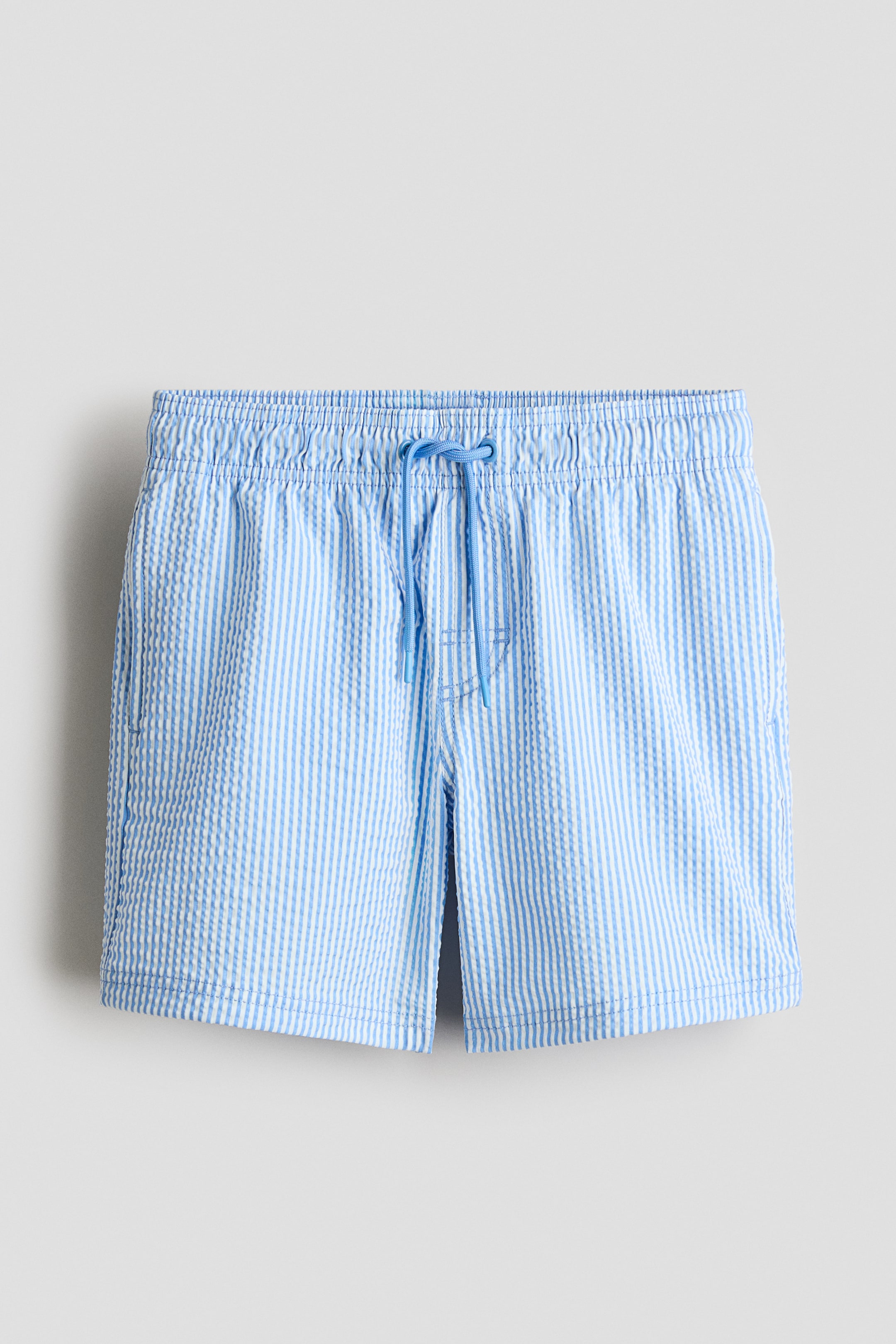 Grotere afbeelding bekijken: De voorkant van de lichtblauw-wit gestreepte seersuckershorts is voorzien van een elastische tailleband met een lichtblauw trekkoord dat in het midden is geknoopt, en zichtbare zijzakken.