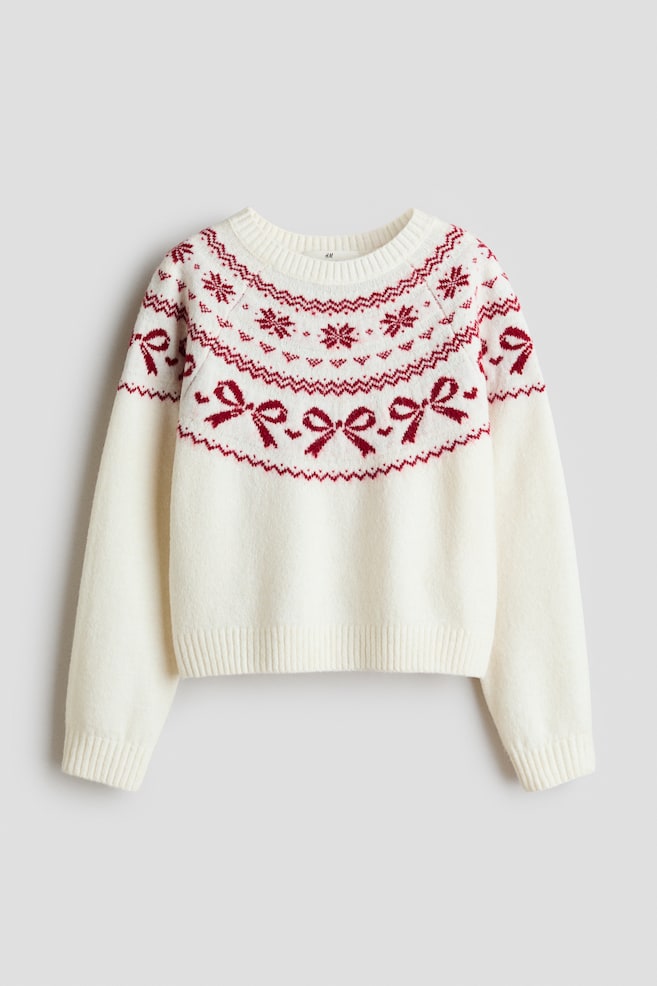 A cream-coloured knit jumper is displayed from the front, featuring a red Fair Isle-style pattern across the yoke, shoulders, and upper sleeves. The design includes rows of red snowflakes, geometric borders, and delicate bows, with the jumper finished with a ribbed crew neck, cuffs, and hem. - 1