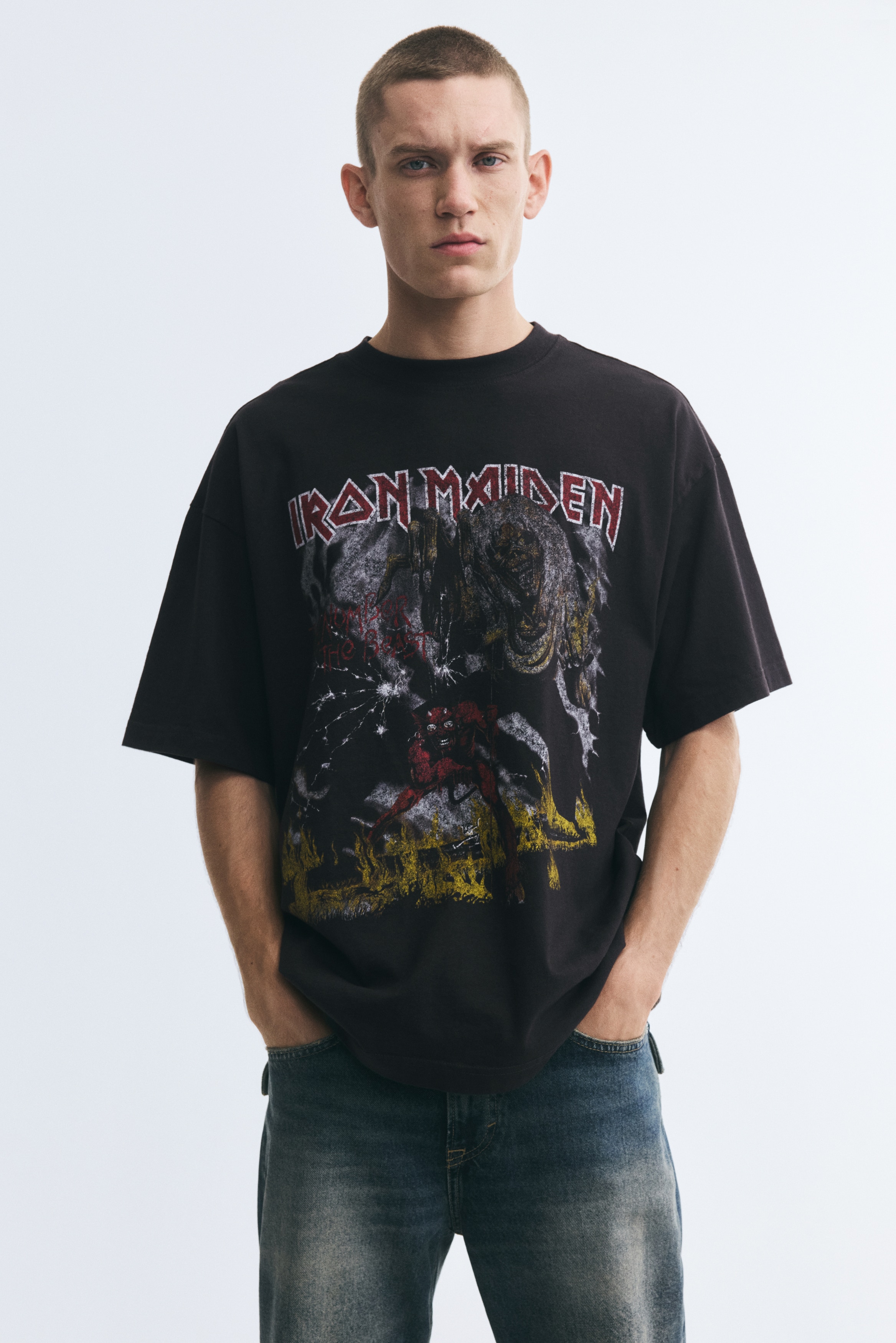A black oversized t-shirt in heavyweight cotton jersey is laid flat, showcasing a prominent graphic print motif on its front. The design features "Iron Maiden" text above a monstrous figure and a red demonic entity, encircled by distressed yellow and white elements. It is crafted with a rib-trimmed neckline, dropped shoulders, and a straight-cut hem, offering a baggy, extra-loose silhouette.