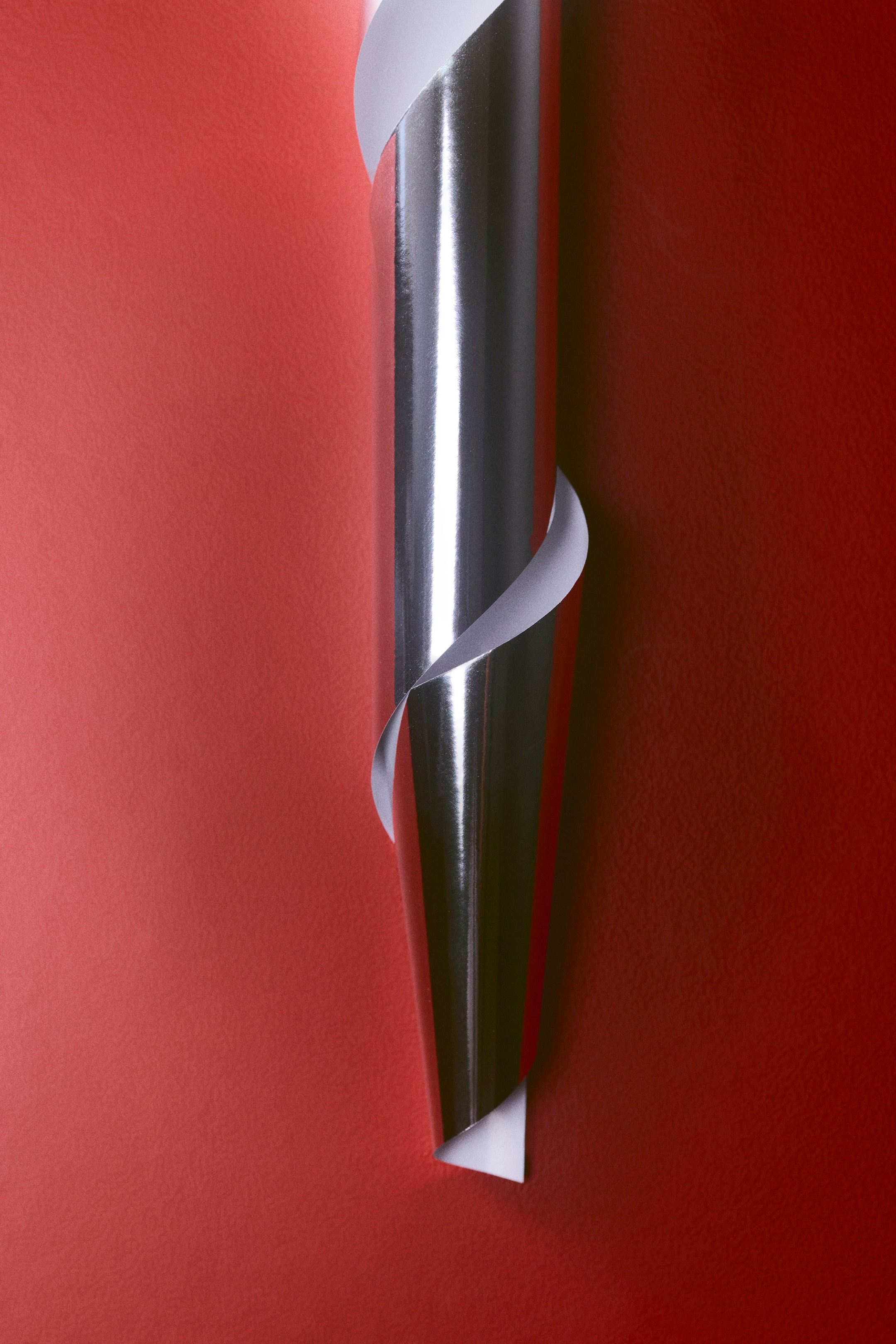 Visualizza immagine più grande: Carta da regalo con un foglio rosso opaco e testurizzato che mostra una spirale verticale di carta da regalo color argento metallizzato. La carta argentata si sta arrotolando per rivelare la sua superficie frontale altamente riflettente e il lato posteriore bianco semplice.