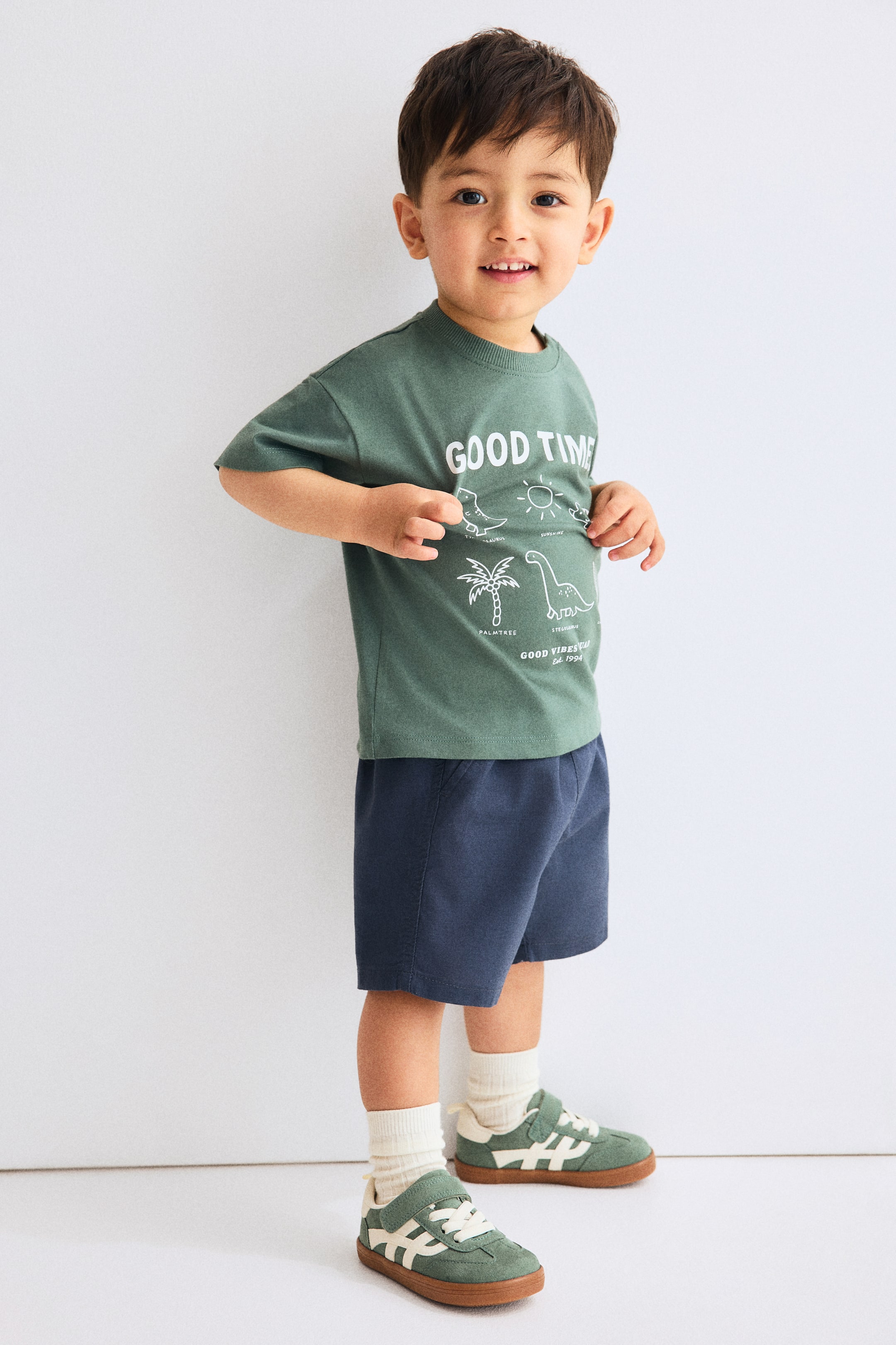 Zobrazit větší obrázek: Zelené tričko s kulatým výstřihem, na kterém jsou bílé obrysové grafiky včetně nápisů "GOOD TIME", slunce, dinosaura, palmy, "GOOD VIBES C." a "Est. 1994", je nošeno s tmavě modrými šortkami s elastickým pasem. Oblečení je prezentováno z mírného úhlu zepředu zleva.