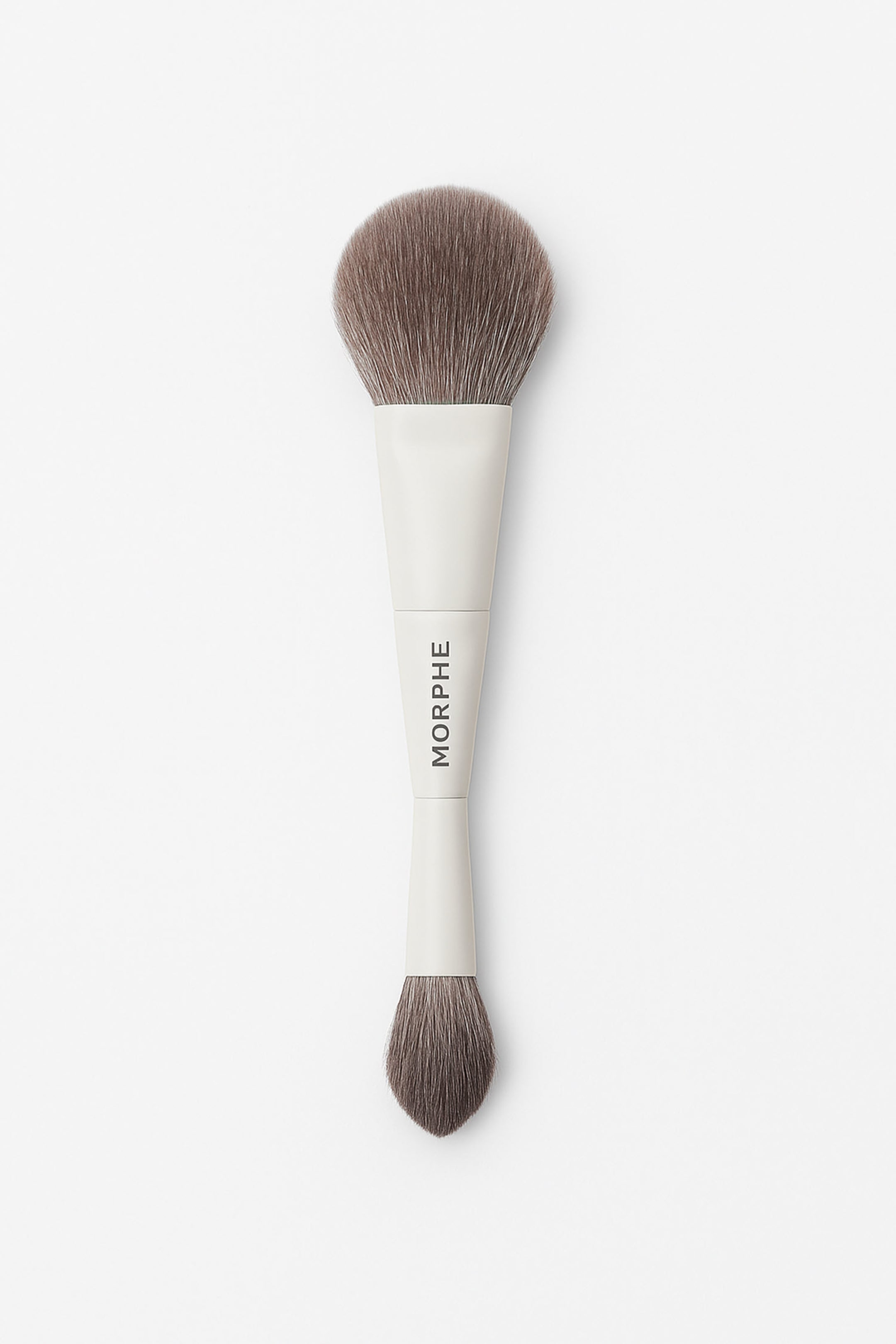 Visa större bild: En dubbeländad makeupborste är orienterad vertikalt och visar ett stort, fluffigt rundat borsthuvud med bruna borststrån upptill. Ett mindre, avsmalnande borsthuvud med bruna borststrån finns nedtill, kopplat med ett ljusfärgat handtag som visar texten "MORPHE" vertikalt i mitten.