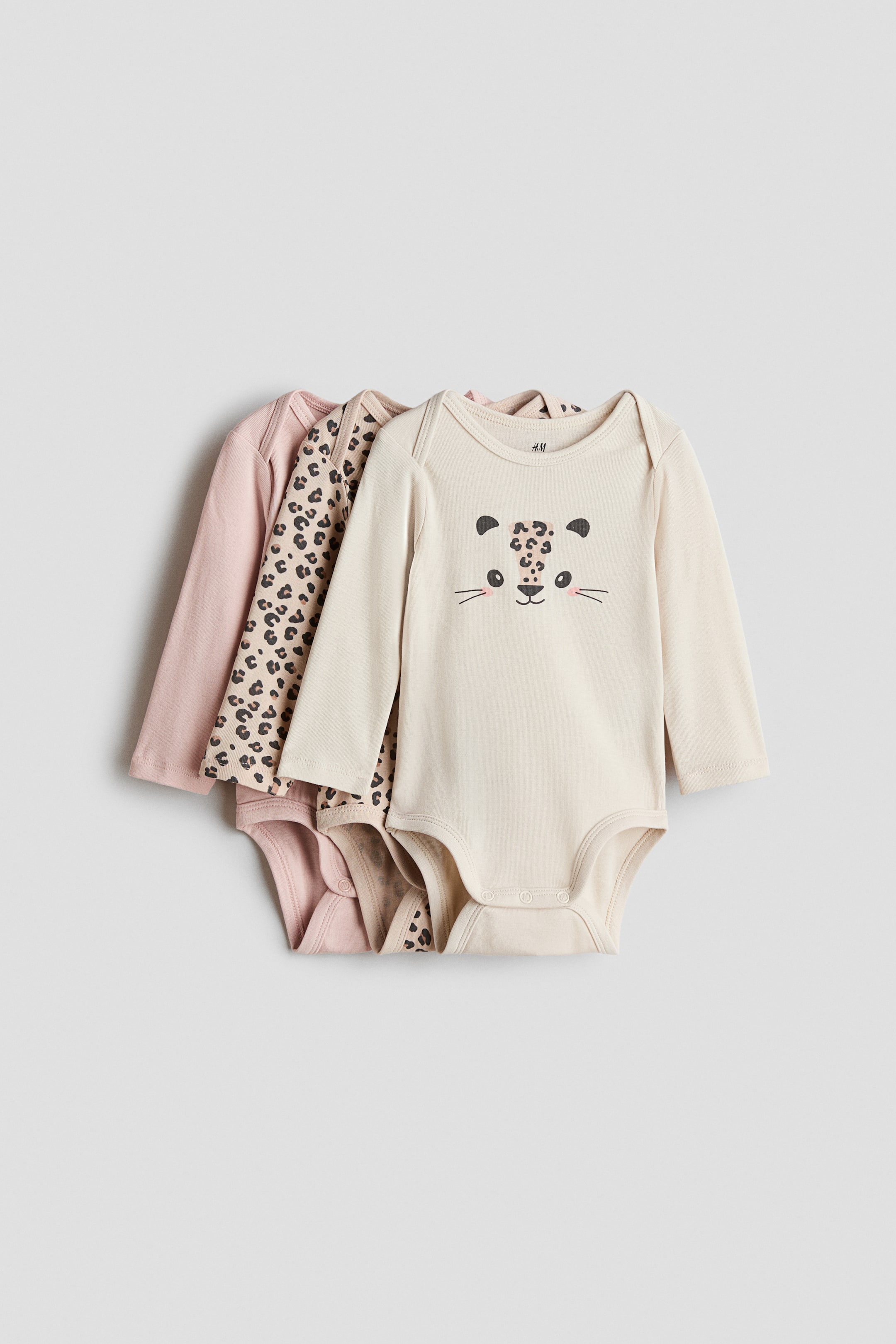 View larger image: Three long-sleeved cotton-jersey bodysuits lie stacked, the front one in light beige featuring a leopard-face graphic print. The bodysuit has an envelope neck and press-studs at the bottom. A second bodysuit with a leopard-spot print and a third solid pink bodysuit are partially visible underneath.