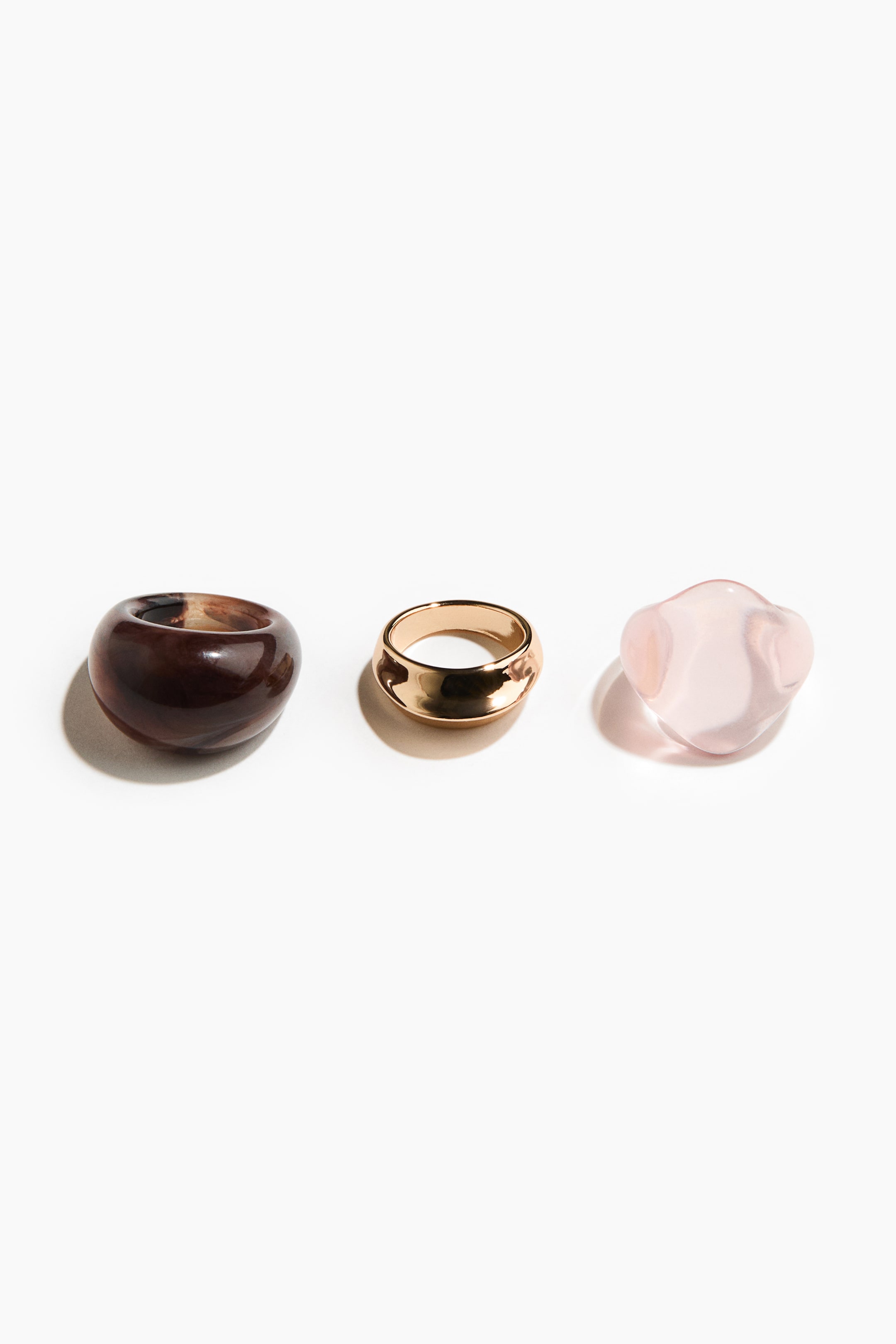 Vis større bilde: De tre ringene er vist frem, hver med et unikt design: en kraftig, mørk brun ring i harpiks med virvlende mønstre som viser frem sitt brede, avrundede bånd, og en bred ring i gullfarget metall med et skulpturert, lett konkavt bånd. Den tredje er en gjennomsiktig, uregelmessig lys rosa steinring med bølgende overflater.