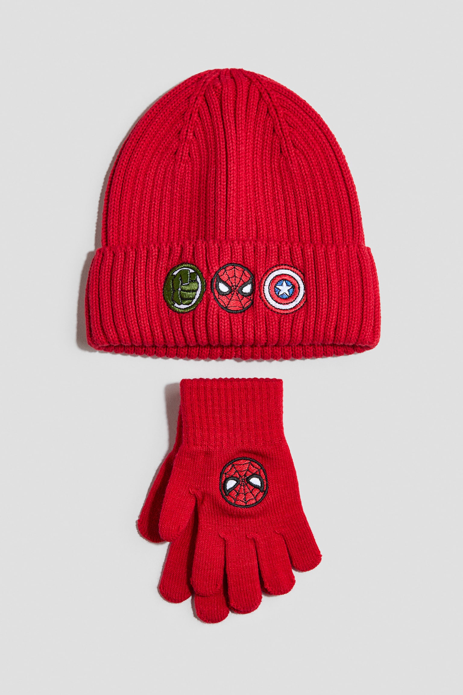 A red ribbed-knit beanie and glove set is laid flat, facing front. The beanie features a folded cuff decorated with three circular embroidered patches of Hulk's fist, Spider-Man's mask, and Captain America's shield, while the matching knit gloves display a Spider-Man mask patch on the back of the hand.