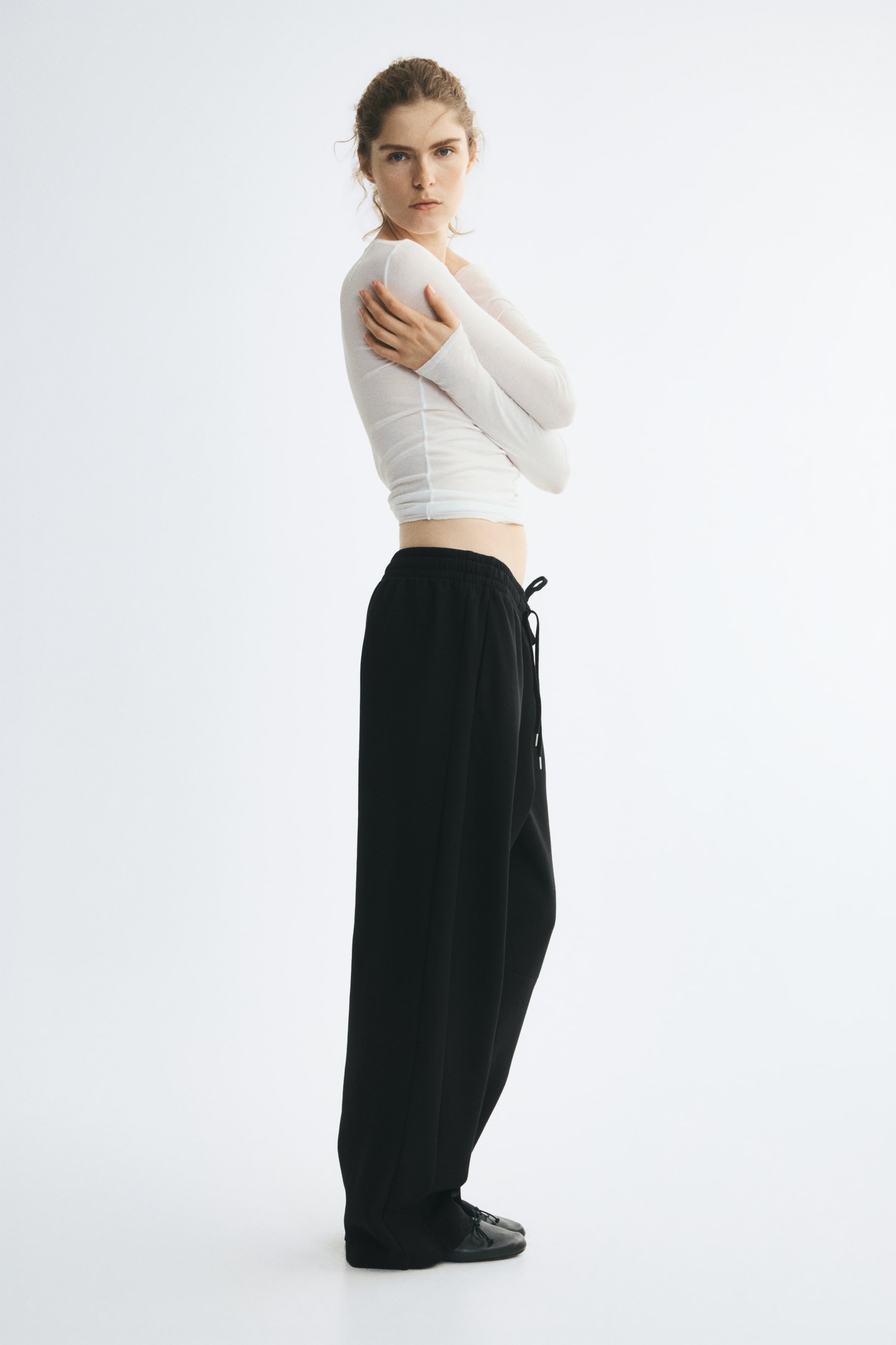 Grösseres Bild ansehen: Schwarze Trackpants in lockerem Schnitt mit hohem, elastischem Bund und Kordelzug werden vom Model getragen. Die Hose ist mit einem transparenten, weissen Langarm-Top kombiniert.