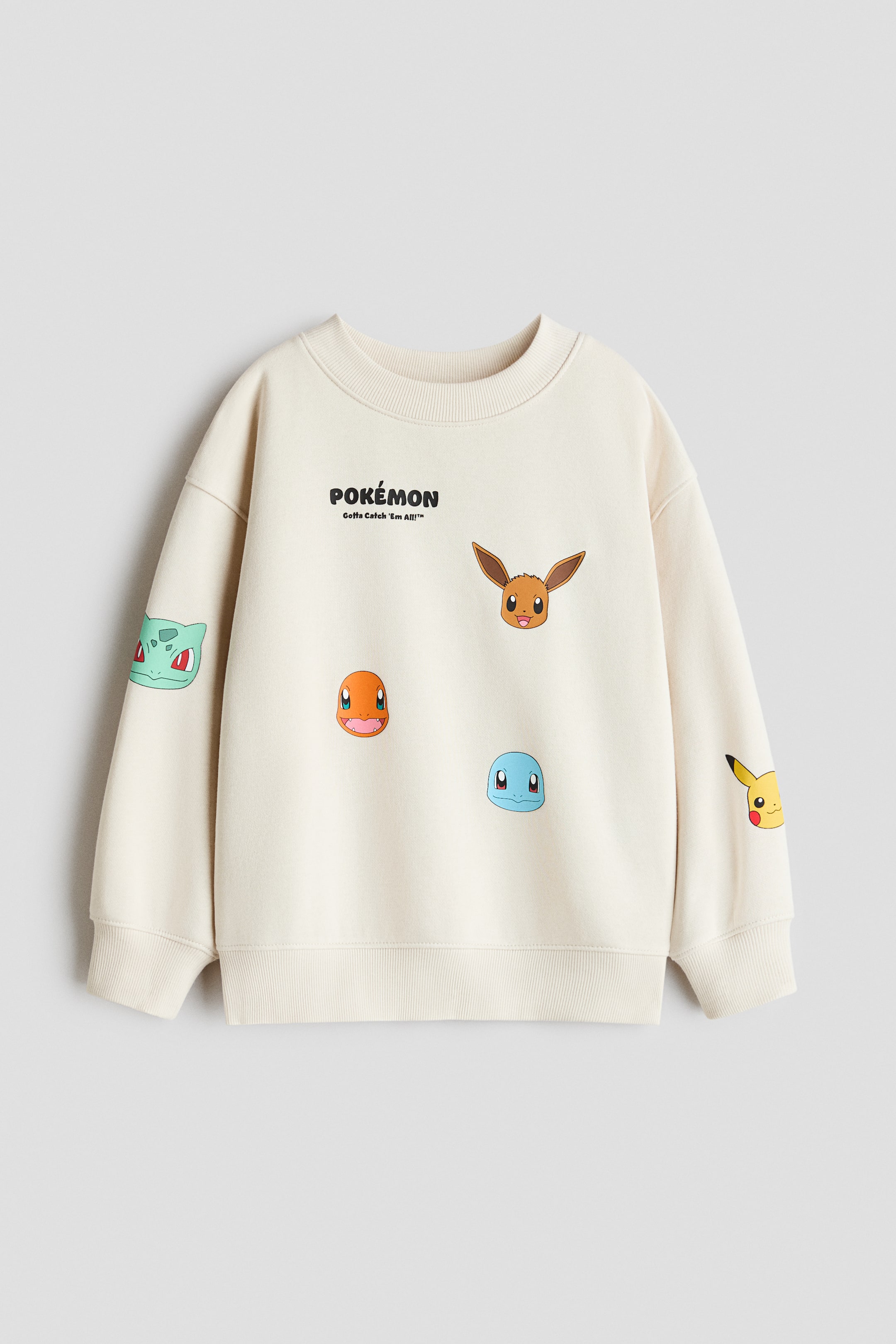 Visa större bild: En krämfärgad sweatshirt med rund, ribbad halsringning, nedhasade axlar och långa ärmar. Ribbstickning finns vid muddar och fåll. Framsidan visar texten "Pokémon Gotta Catch 'Em All!" med tryckta ansikten av Eevee, Charmander och Squirtle, medan fler karaktärsansikten, inklusive Bulbasaur och Pikachu, syns på ärmarna.
