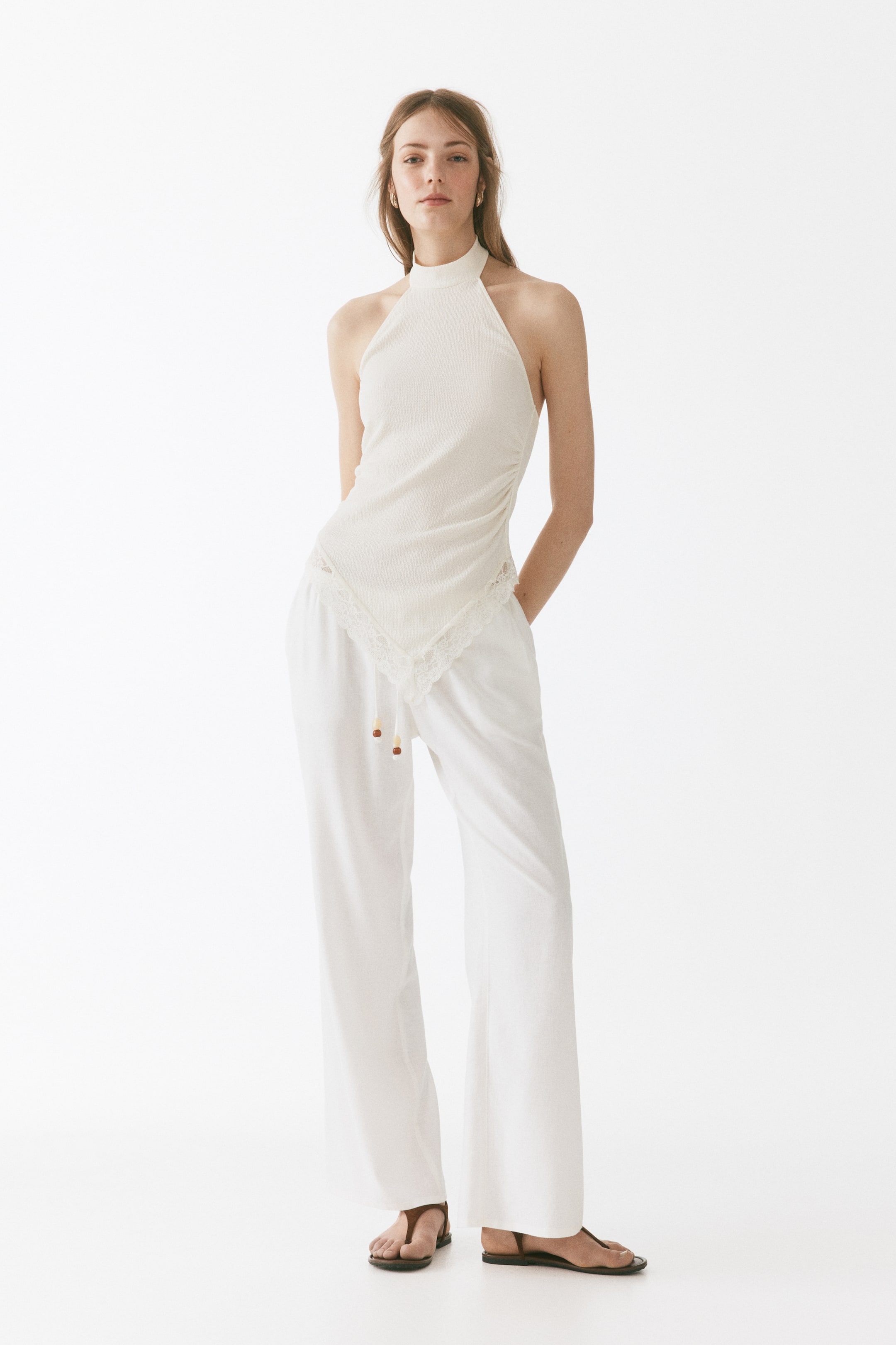 Ver imagen más grande: Se presenta un top blanco de canalé con cuello halter y un cuello alto simulado, con un dobladillo asimétrico adornado con encaje, combinado con pantalones blancos de pierna ancha que cuentan con una cintura de cordón con cuentas de madera.