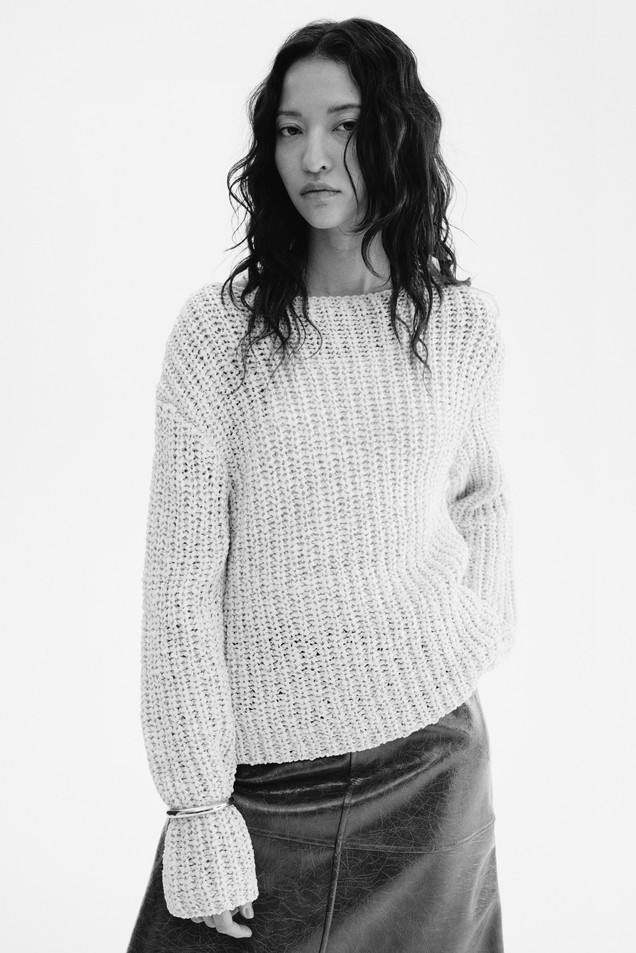 View larger image: The model is seen wearing a jumper crafted in a textured rib knit, featuring a boat neckline, dropped shoulders, and long sleeves. She casually tucks one hand into the front of the jumper, while a silver-toned bracelet adorns one of the sleeves. The jumper is elegantly paired with a dark, textured skirt.