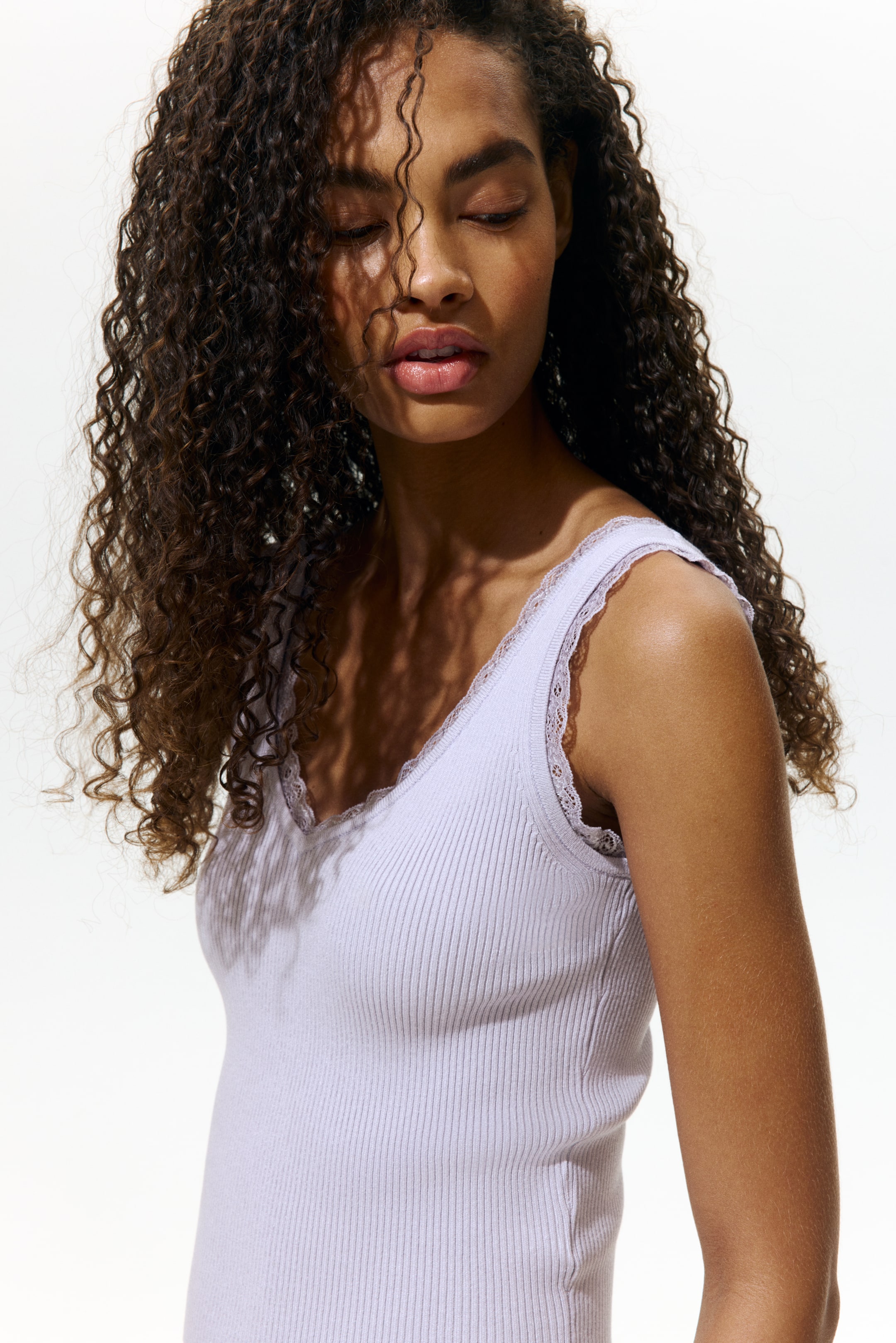 Größeres Bild ansehen: Ein figurbetontes, helllila geripptes Trägertop wird vom Model getragen. Es ist mit einem V-Ausschnitt gestaltet und mit Spitze an Ausschnitt und Armlöchern verziert.