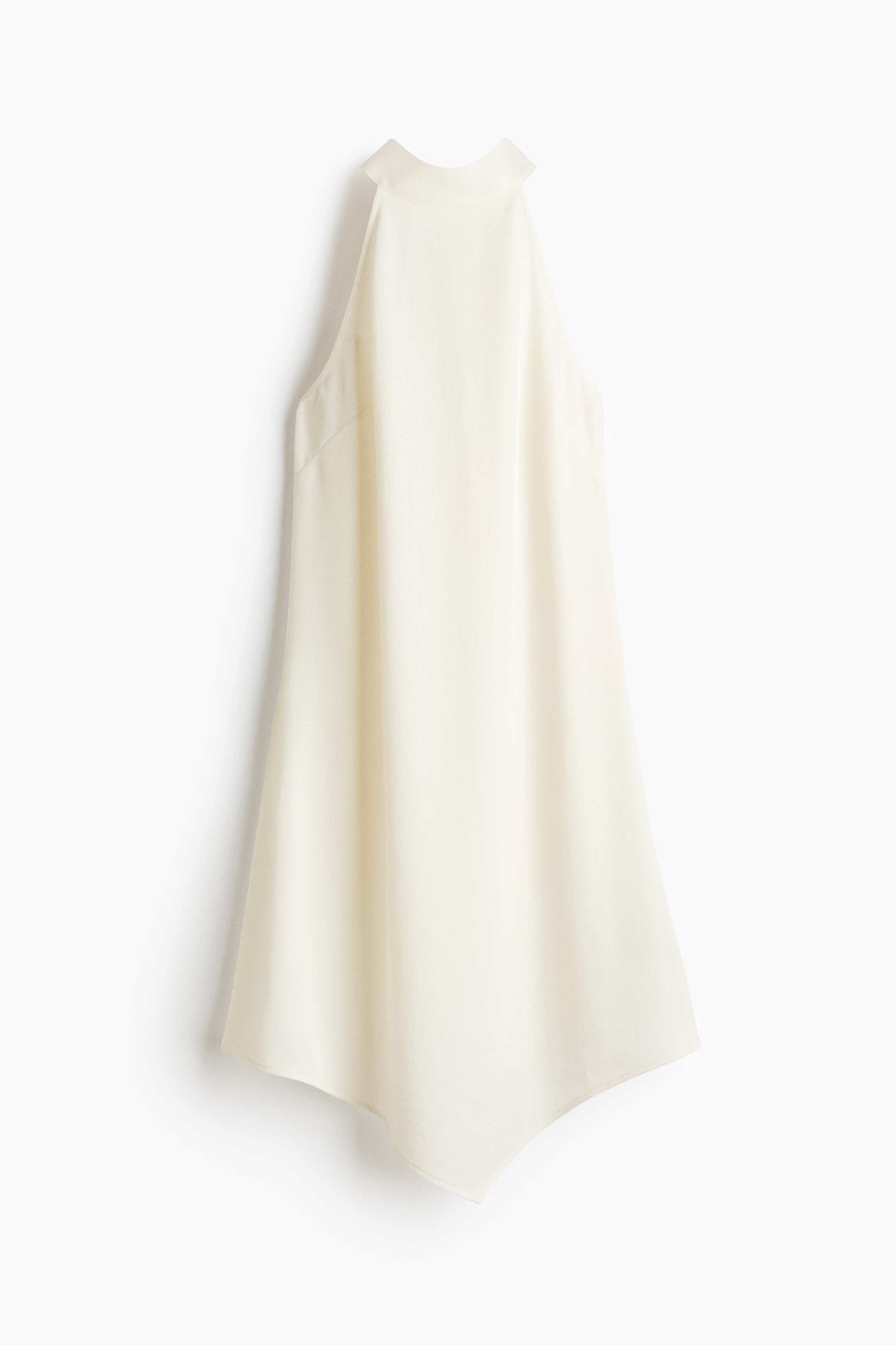 View larger image: An off-white halter top is hanging, showcasing a high neck that forms a standing collar at the back. It features a loose, flowing silhouette and is completed with an asymmetrical hemline, creating soft points at the sides.