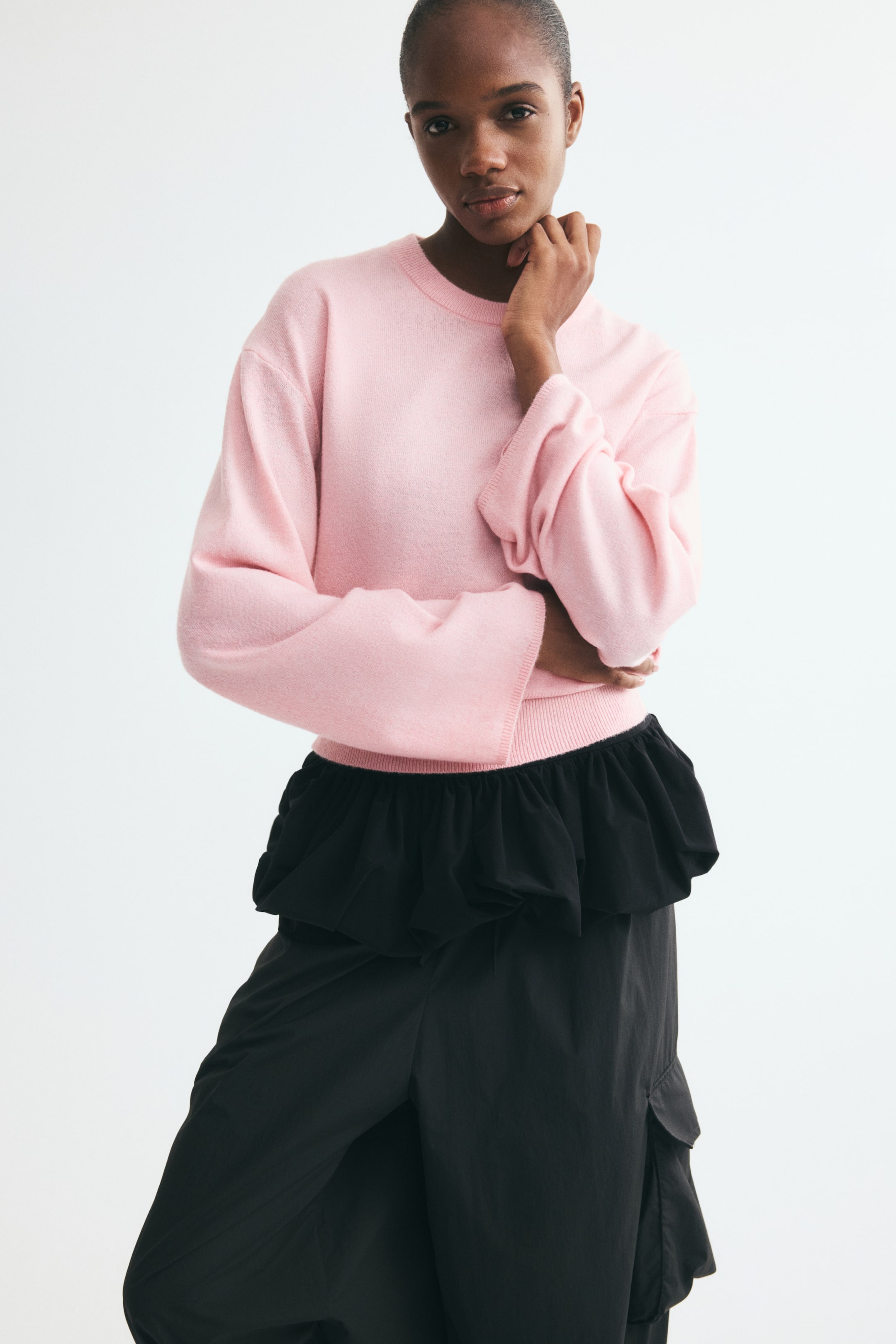 Visualizza l'immagine ingrandita: Un maglione in maglia rosa chiaro, corto, con scollo rotondo, spalle scese e ampio orlo a coste, indossato dalla modella. Il maglione è abbinato a un capo nero con un evidente punto vita arricciato e completato da pantaloni neri.