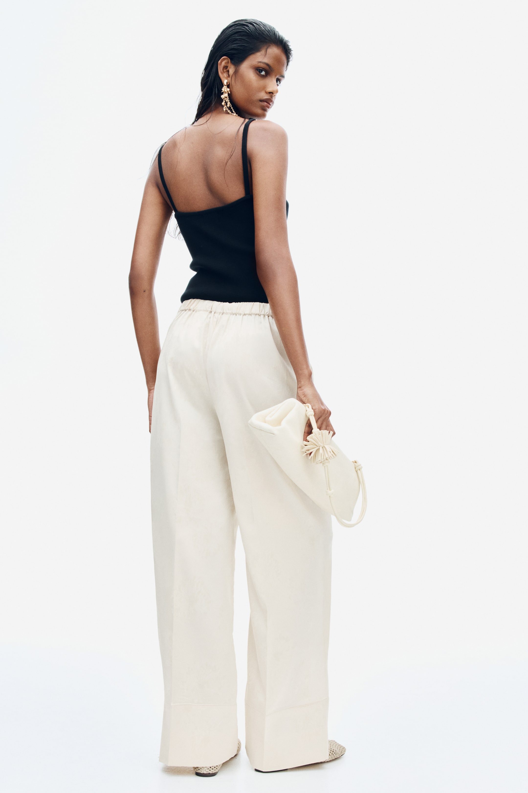View larger image: Cream-coloured, loose-fit pants in jacquard-weave satin are being worn by the model, showcasing a high waist with an elasticated back waistband and wide legs with pressed creases. The pants are styled with a tucked-in black tank top, and the model is holding a coordinating cream clutch bag.