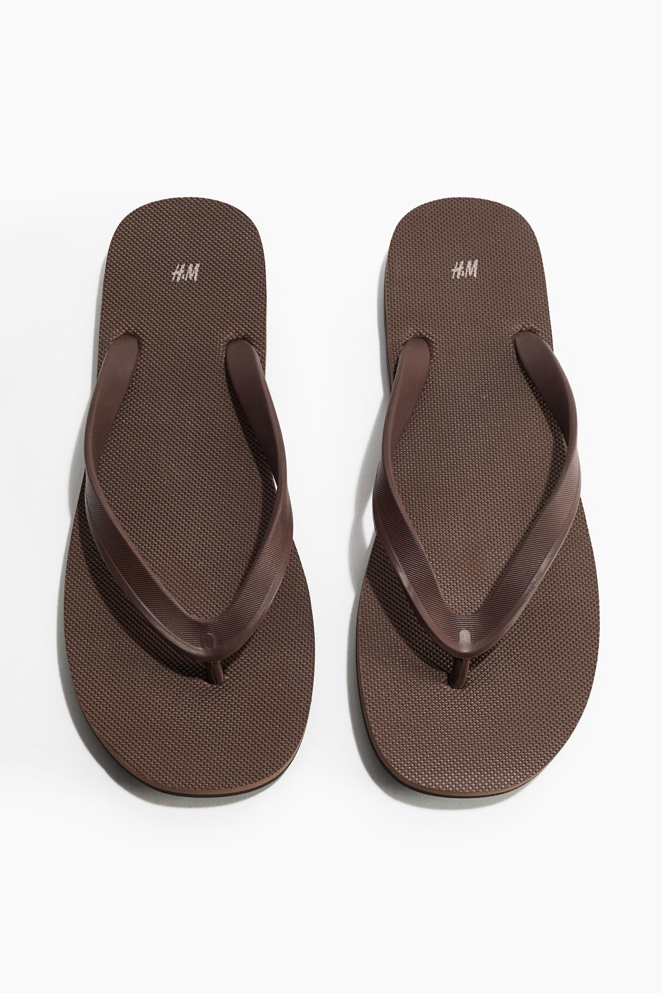 Ver imagem maior: Um par de chinelos de borracha castanhos apresenta uma palmilha texturizada com o logótipo da H&M, exibindo tiras castanhas a condizer e um fino separador de dedos.