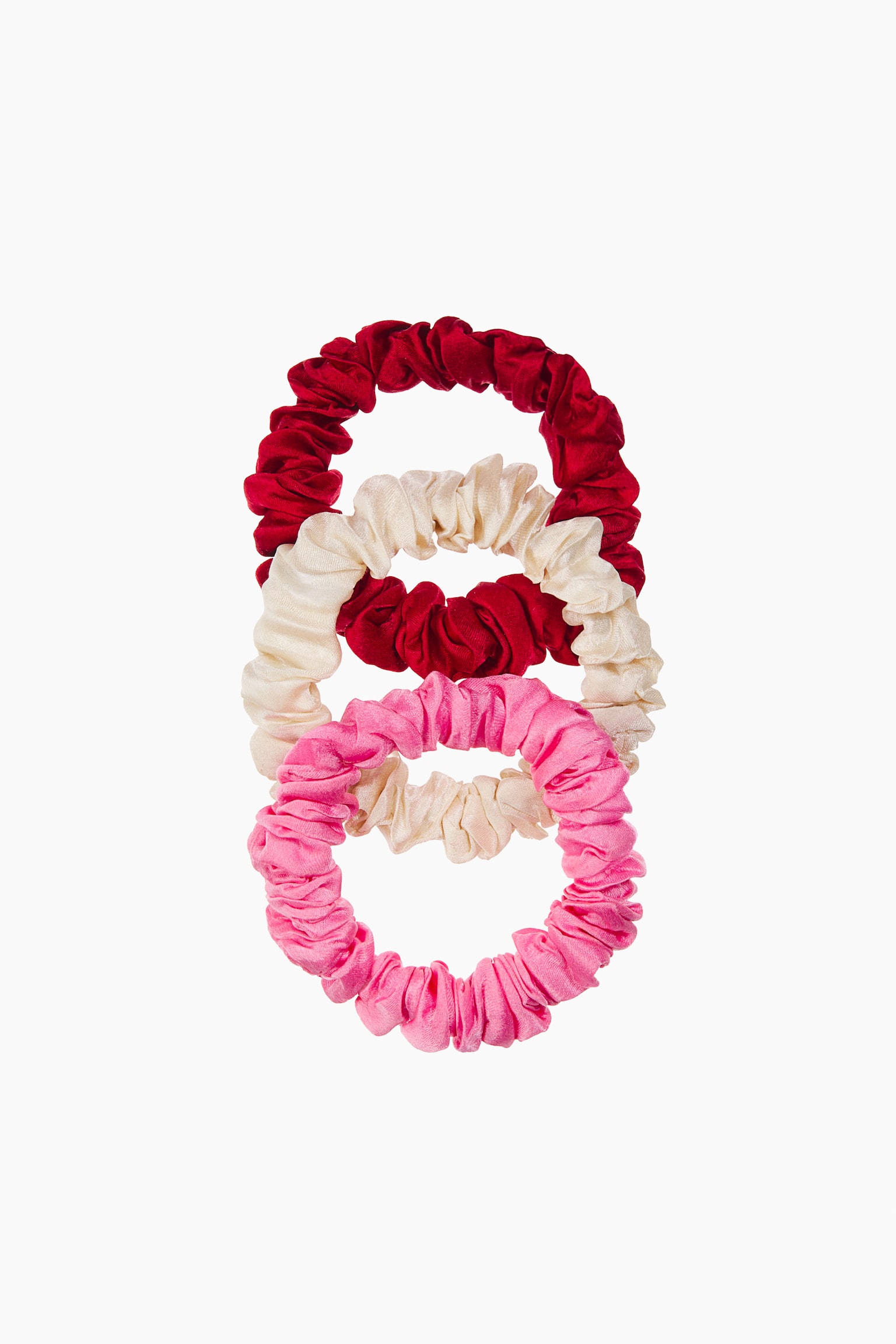 Tre elastiske scrunchies i rød, creme og pink fremstår i en lodret, overlappende stak. Hver scrunchie har et rynket, draperet design i glat, skinnende silkestof, vist forfra for at fremhæve dens elastiske struktur.