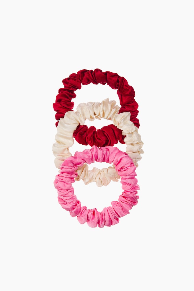 Trois chouchous élastiqués, en rouge, crème et rose, apparaissent en pile verticale superposée. Chaque chouchou présente un design froncé et plissé, confectionné en soie lisse et lustrée, vu de face pour mettre en valeur sa structure élastiquée. - 1