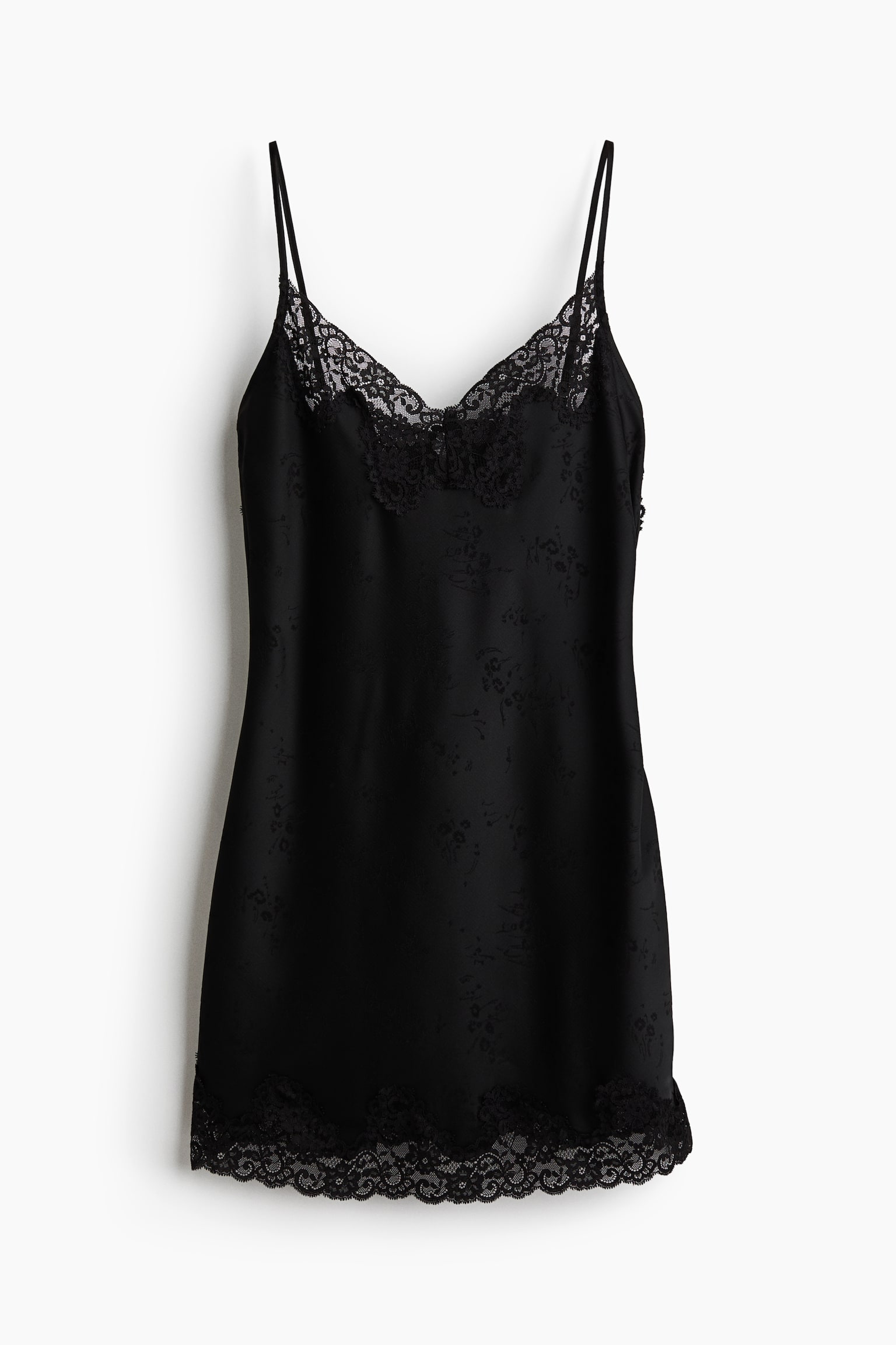 Un abito sottoveste nero è appeso in vista frontale, caratterizzato da un discreto motivo floreale jacquard tono su tono su tutta la superficie. Il design include sottili spalline spaghetti, uno scollo a V rifinito con delicato pizzo nero e un bordo in pizzo smerlato coordinato lungo lâorlo.