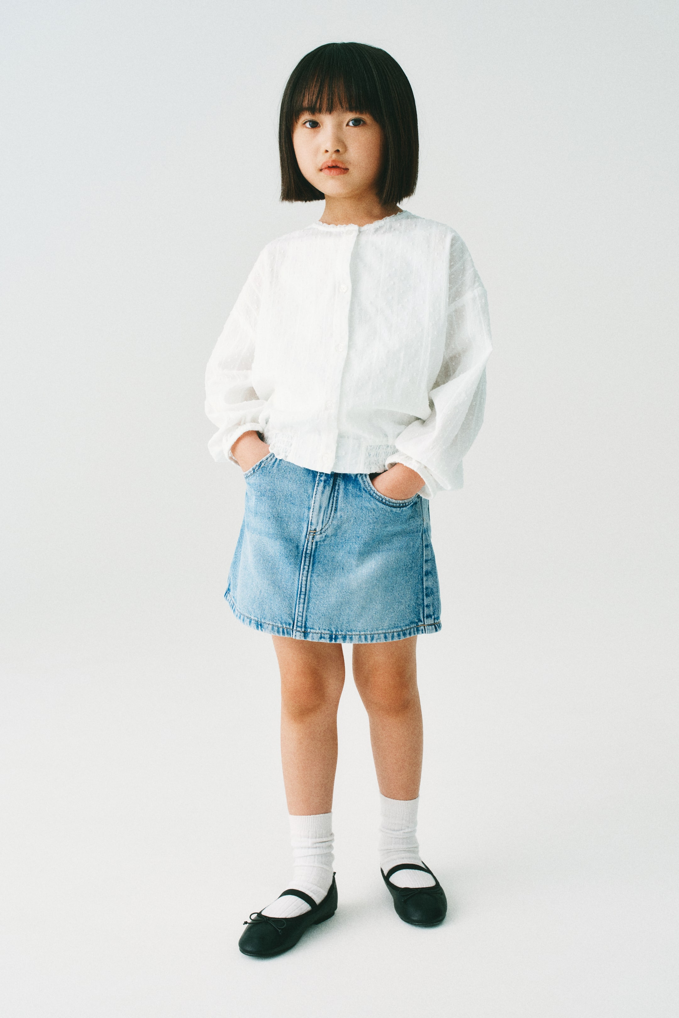 拡大画像を表示: モデルが着用しているのは、ルーズフィットの白いコットンプルメティブラウスです。丸いレーストリムのネックライン、前面のボタン、ドロップショルダー、長いバルーンスリーブには伸縮性のあるカフスが施され、裾には小さなフリルトリム付きの伸縮性のあるスモックが特徴です。このブラウスはデニムスカートとスタイリングされています。