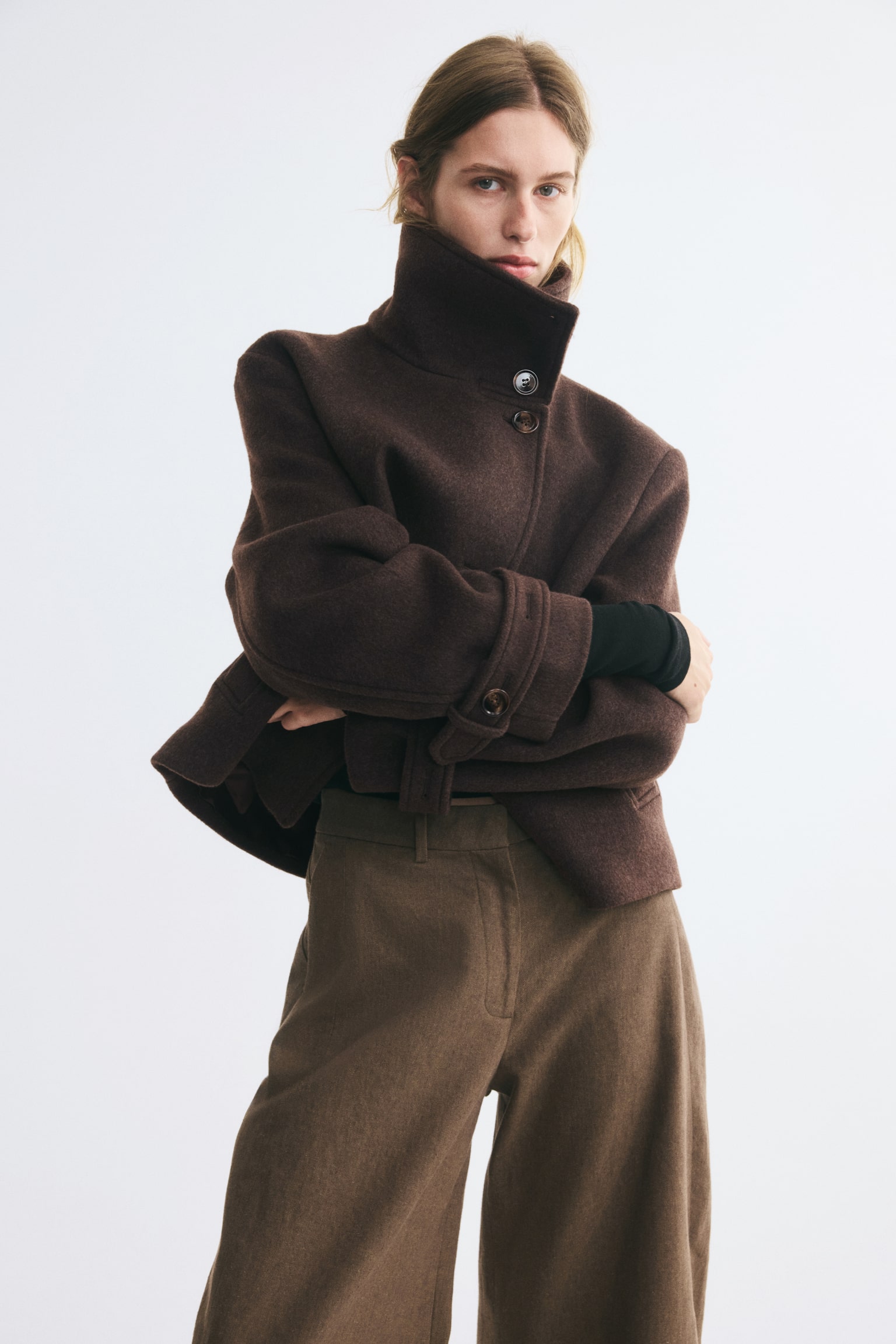 A dark brown wool jacket is facing front, showcasing a cropped, boxy silhouette with an asymmetrical button-front closure. It is featuring a high, buttoned stand-up collar, vertical welt pockets, and wide sleeves with buttoned cuff tabs. - 1