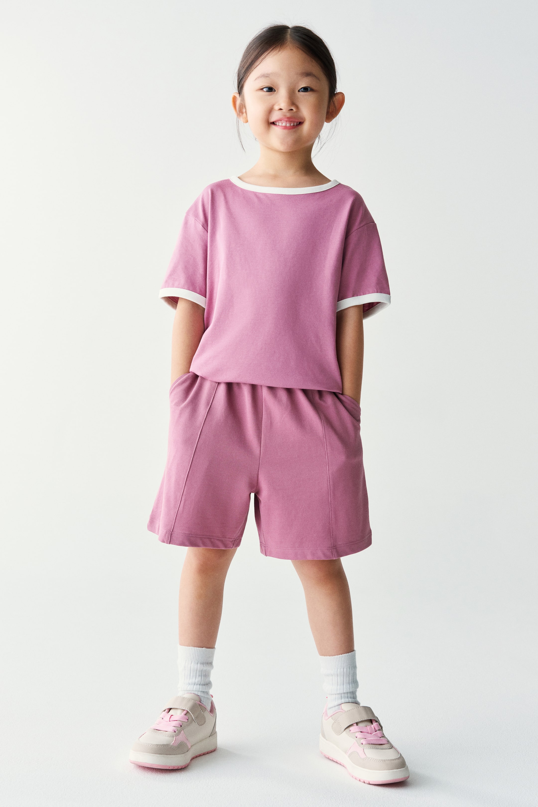 View larger image: A mauve pink two-piece set, consisting of a short-sleeved T-shirt with white trim at the collar and cuffs and matching shorts with front seam details, is worn by the model. The model is standing with hands resting in the shorts’ pockets, styled with white ribbed socks and beige sneakers.