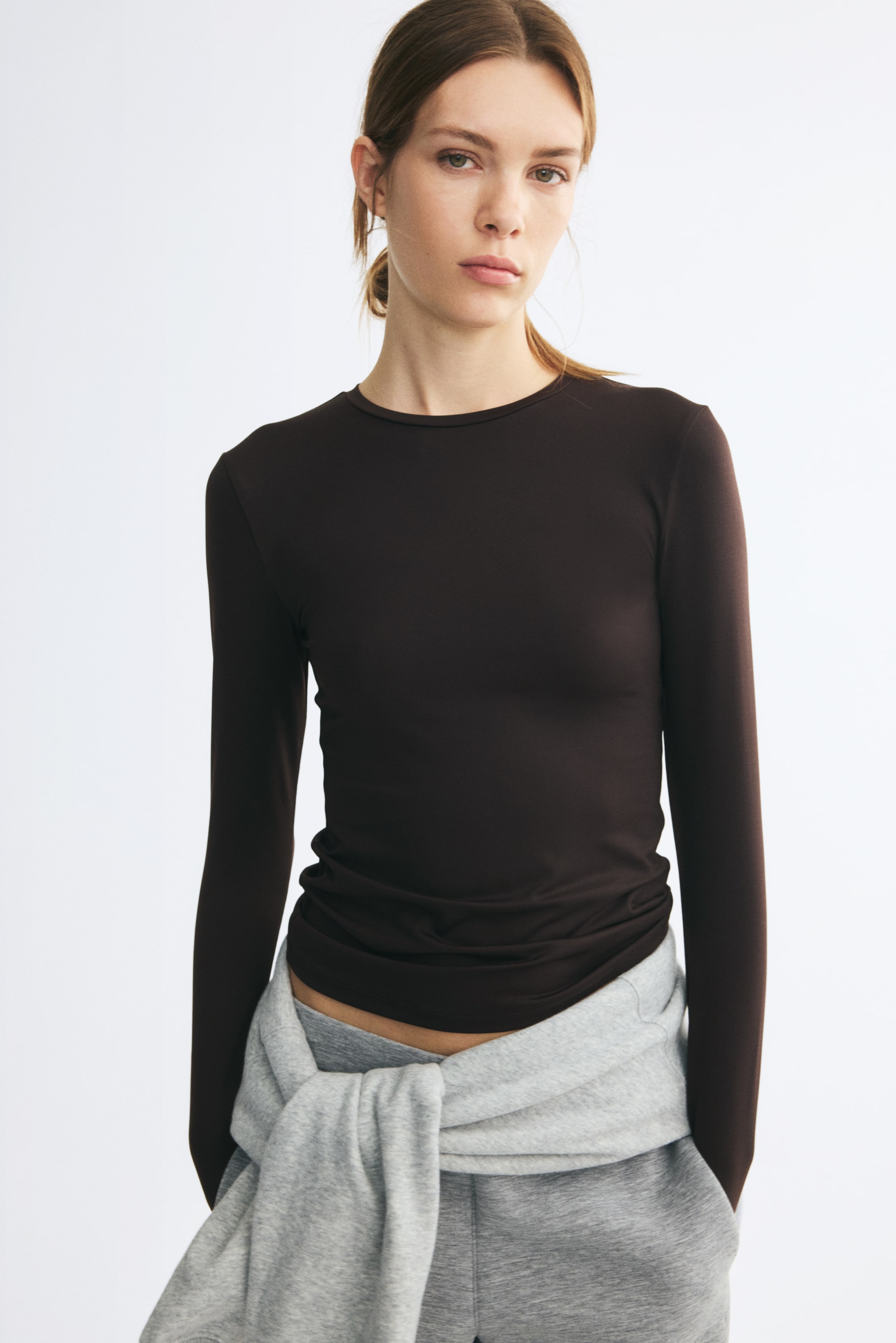 Grotere afbeelding bekijken: Het model draagt een donkerbruine, aansluitende jersey top met een ronde hals met smalle bies en lange mouwen. De top is gestyled met een grijze joggingbroek en een lichtgrijze sweatshirt die om de taille is geknoopt.