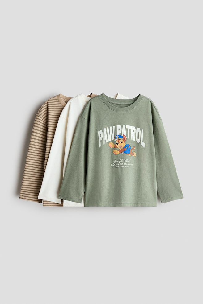 Tres camisetas de algodón de manga larga se superponen y están orientadas hacia delante, mostrando en primer plano una camiseta verde salvia con estampado gráfico de Paw Patrol y cuello redondo con ribete acanalado. Detrás, se aprecian una camiseta lisa en color blanco roto y una camiseta marrón y beige con rayas horizontales. - 1