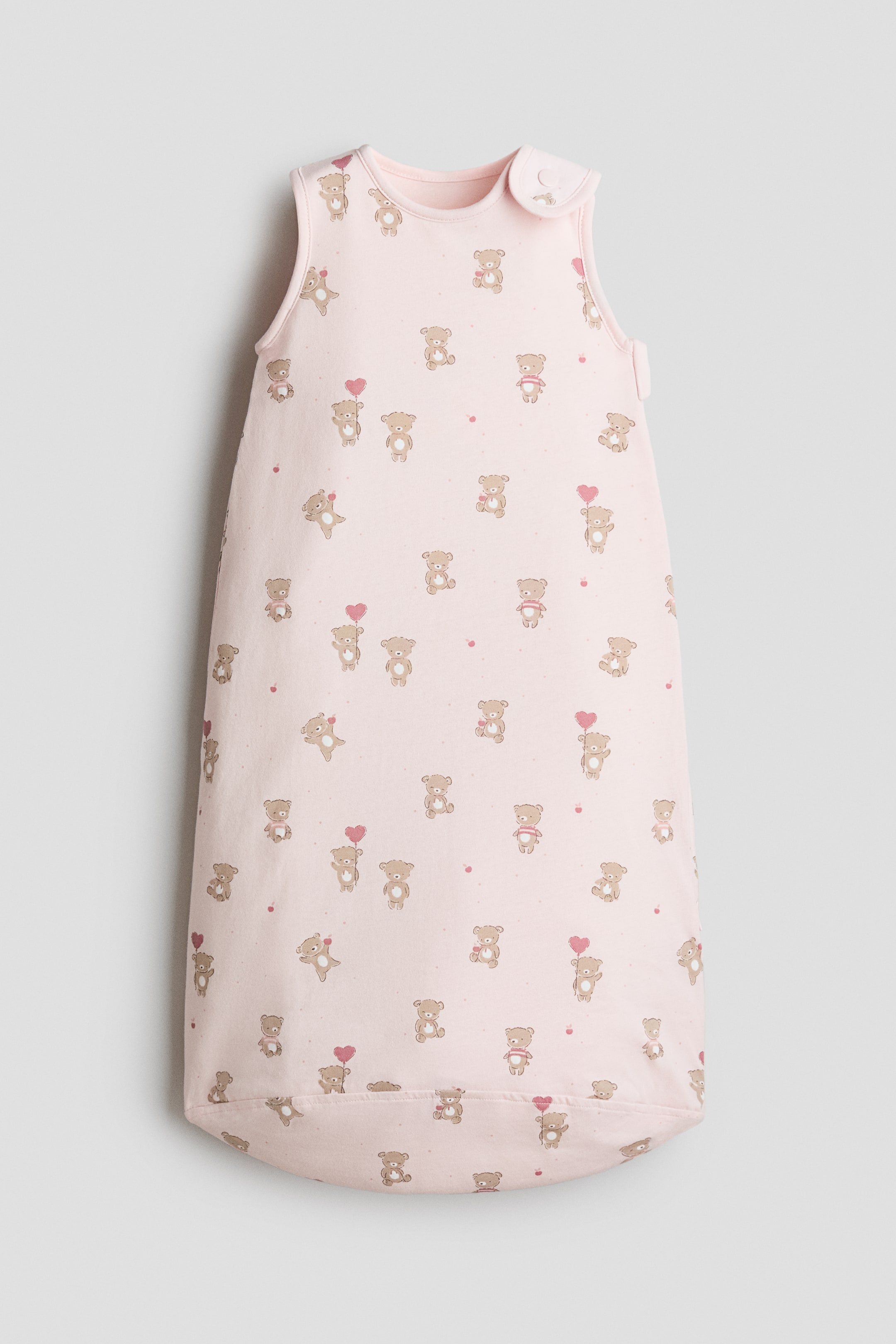 Visualizza immagine più grande: Un sacco nanna imbottito è mostrato frontalmente, caratterizzato da un tessuto jersey stampato rosa chiaro con motivo all-over di orsetti marroni e cuori rosa, e presenta una chiusura a pressione su una spalla e una cerniera verticale lungo il lato destro.