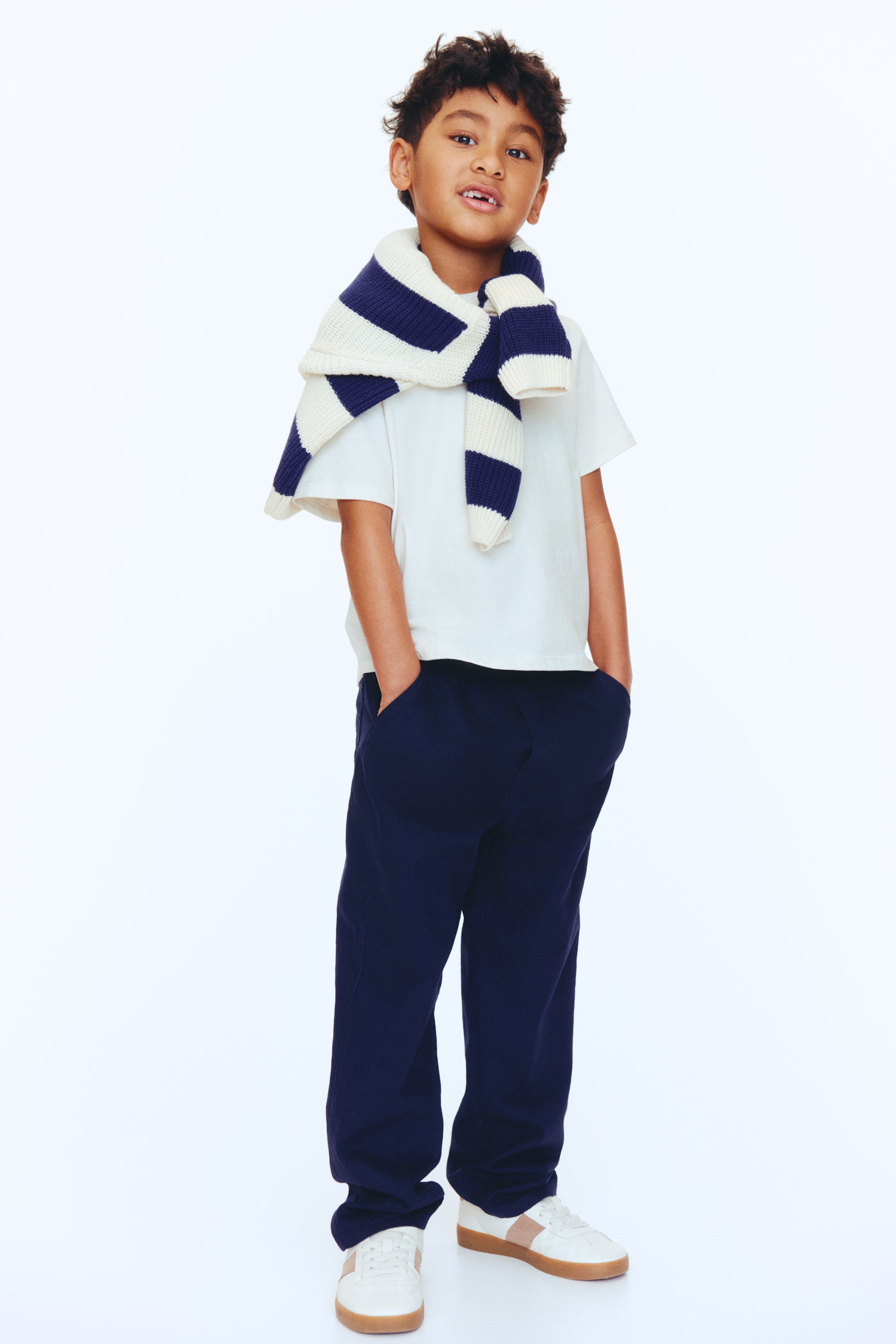 Visa större bild: Ett barnklädeset består av en vit t-shirt med rund hals, marinblå byxor med sidfickor och vita sneakers med bruna sidostripes och gummisulor, med en vit och marinblå randig stickad tröja knuten runt axlarna.