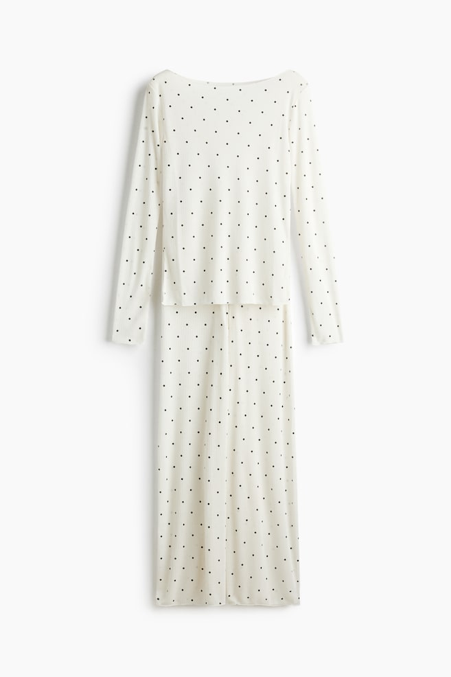 This cream-colored pyjama set is facing front, featuring a long-sleeved top with a boat neckline and matching wide-leg pants in a ribbed jersey fabric with a black polka dot pattern. Both pieces are showing overlocked edges and a slightly sheer texture. - 1