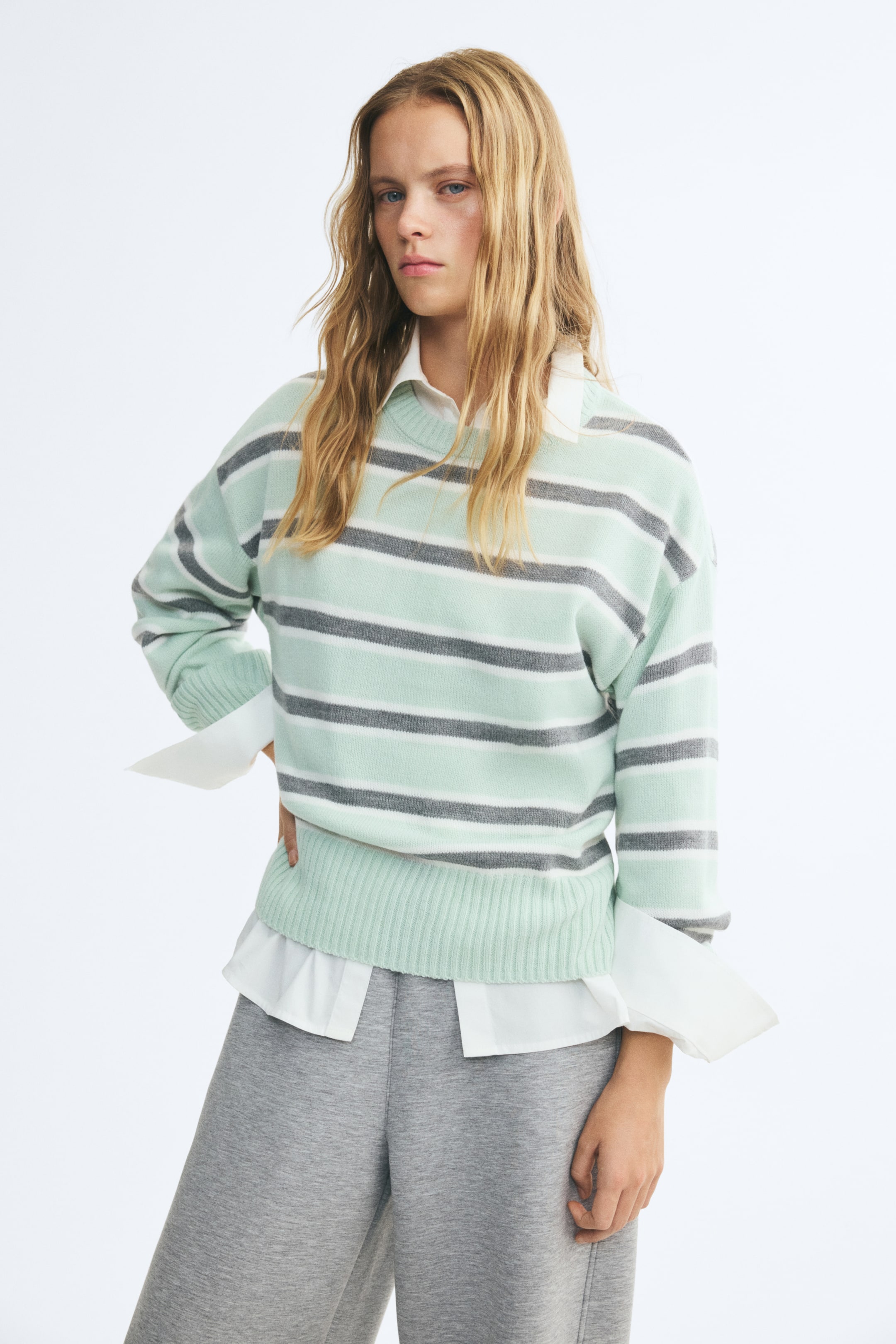 View larger image: A relaxed-fit knit sweater with mint green, white, and gray horizontal stripes is being worn by the model. The sweater features a ribbed crew neck, dropped shoulders, and a ribbed hem, and is layered over a white collared shirt and paired with gray trousers.