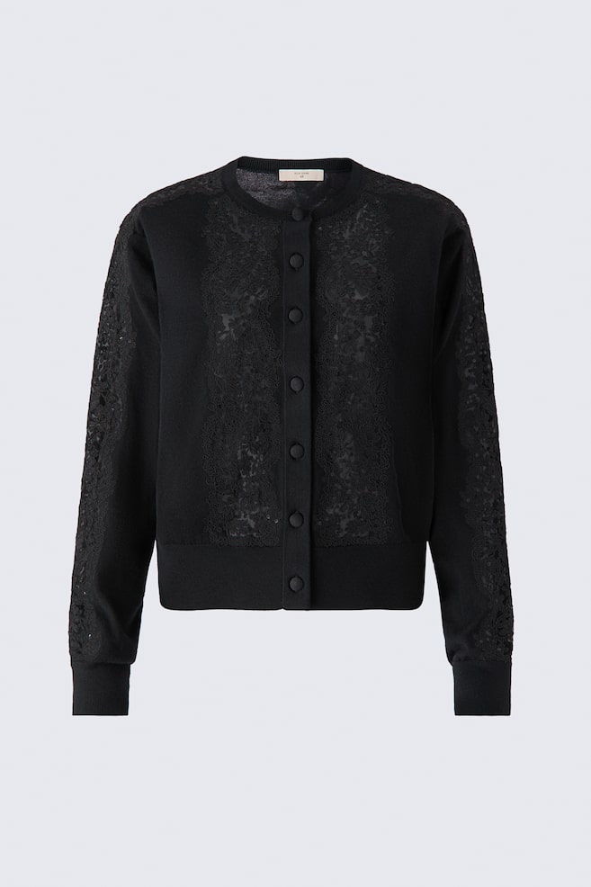 A front-facing studio shot displays a short black knit cardigan featuring a round, rib-trimmed neckline and fabric-covered buttons down the front. Matching black lace panels detailed with tonal sequins are running down both sides of the front placket and along the long sleeves, which are ending in ribbed cuffs and a ribbed hem. - 1
