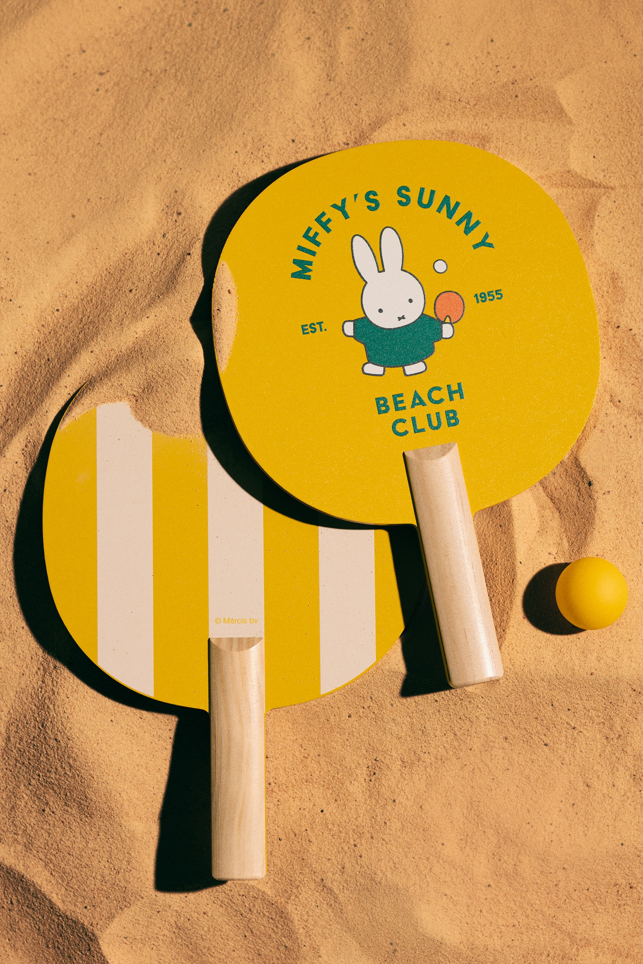 Agrandir l'image: Le set de beach tennis Miffy's Sunny Beach Club présente deux raquettes arrondies et une petite balle jaune, avec les raquettes légèrement superposées. Une raquette est entièrement jaune, arborant un motif de Miffy et du texte, tandis que l'autre est rayée jaune et blanc. Les deux raquettes possèdent des poignées en bois clair, et la balle jaune repose à côté de l'une des poignées.
