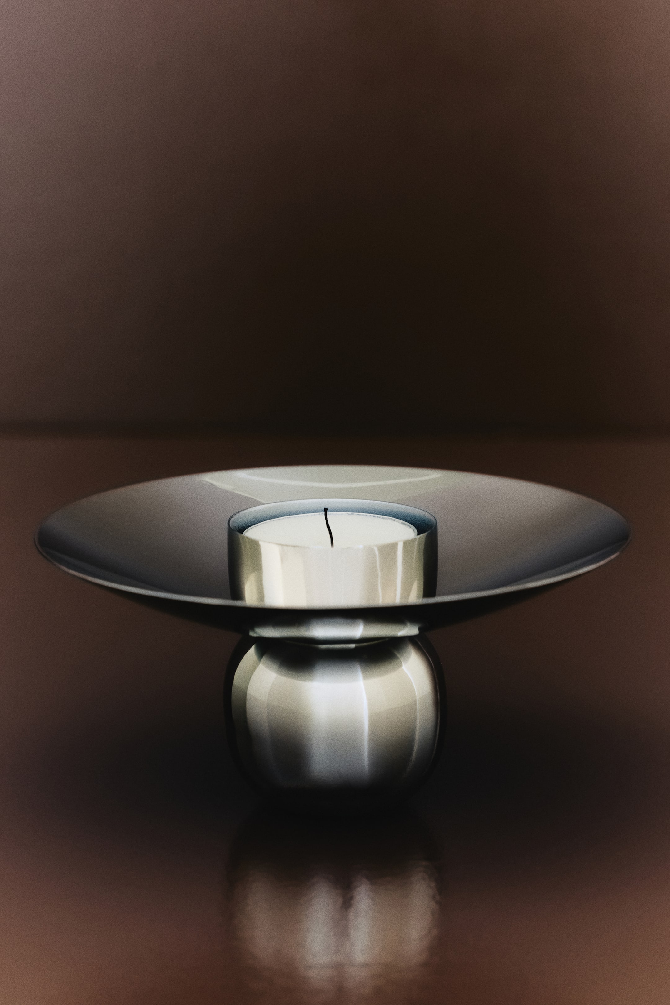 View larger image: The stainless steel tealight holder showcases an elegant design when viewed from the front. It boasts a broad, shallow, concave top surface and a spherical base, with a white tealight candle positioned centrally on the top surface.