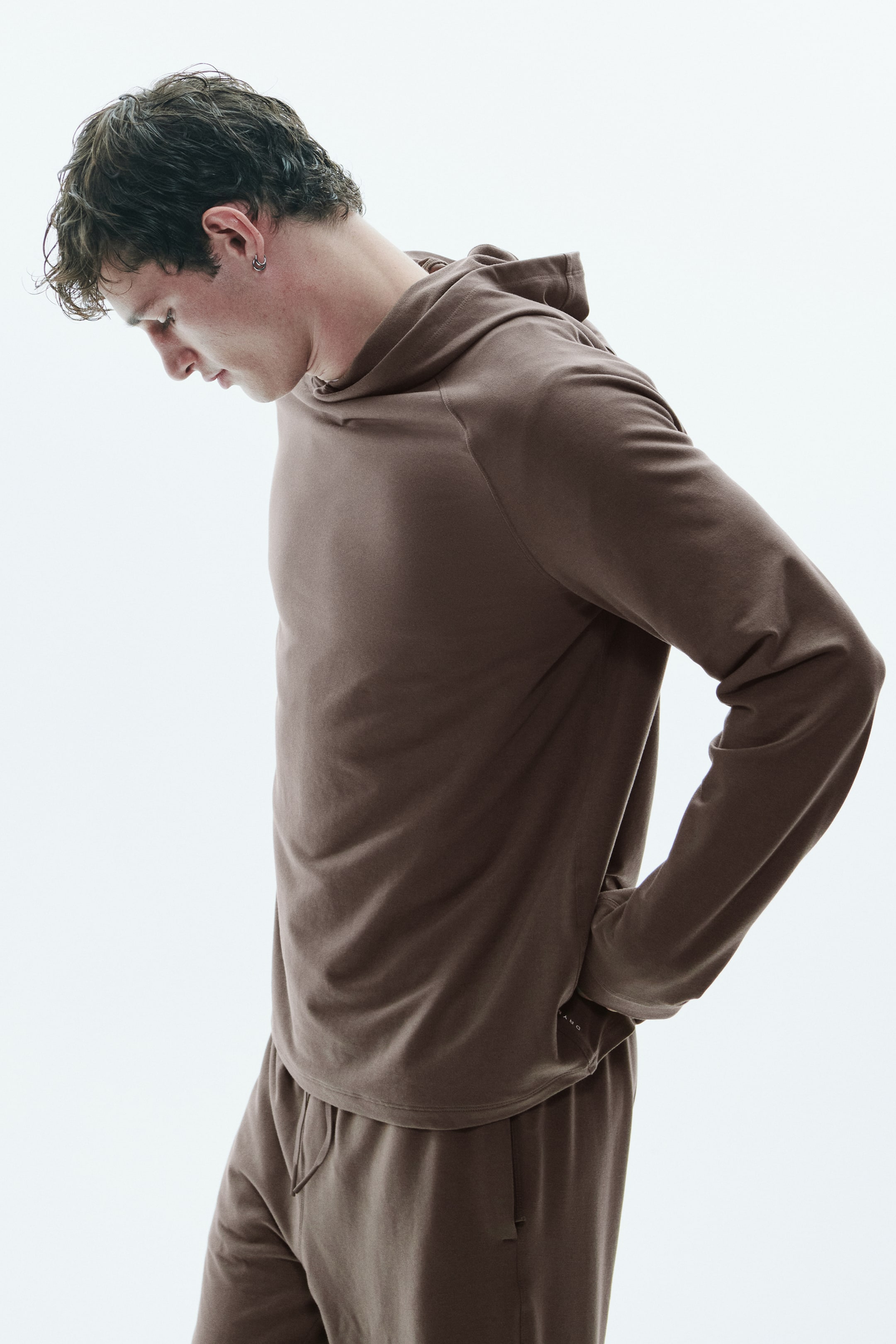 Agrandir l'image: Un sweat à capuche marron léger, doté de manches raglan et d’un ourlet arrondi, est porté par le mannequin. Le sweat à capuche est assorti à un bas à cordon de serrage coordonné, tandis que le mannequin se tient de profil et regarde vers le bas.
