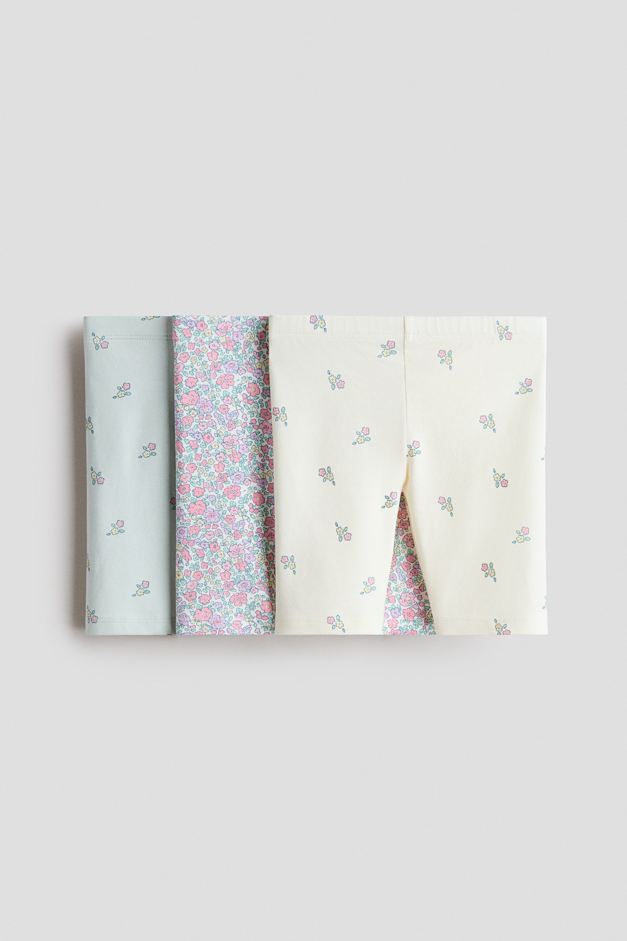 View larger image: Three pairs of children's leggings are placed flat, folded, and arranged side-by-side, showing their top halves. The pair on the left displays a light blue base with scattered small floral patterns. The middle pair features a dense all-over floral print in pink and purple tones. The pair on the right showcases a cream base with scattered small floral patterns.
