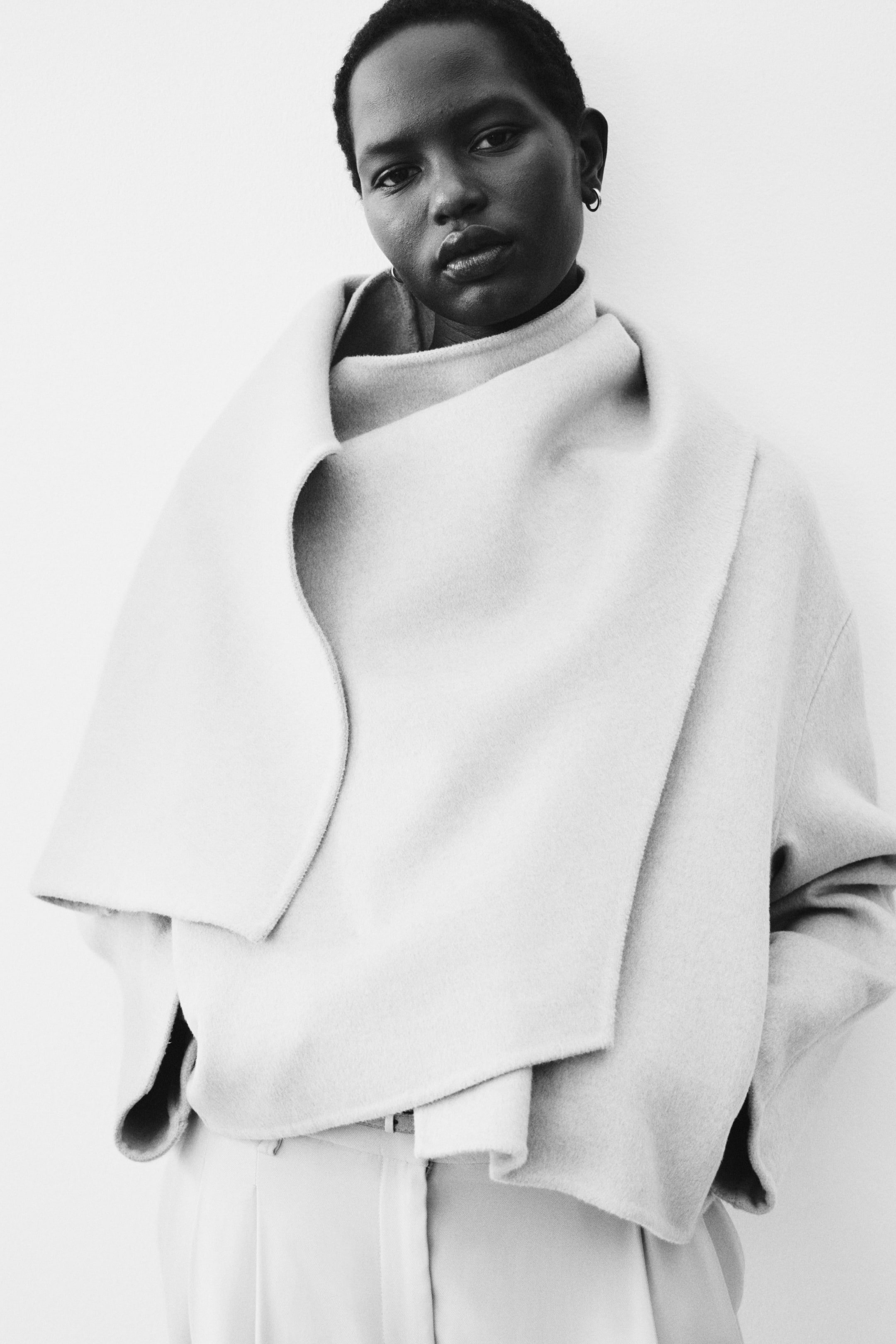 Ingrandisci l'immagine: Un cappotto avvolgente di tonalità chiara presenta un ampio colletto drappeggiato asimmetrico che si curva attorno al collo e al petto. È caratterizzato da maniche larghe ed è abbinato a un top a collo alto e pantaloni coordinati.